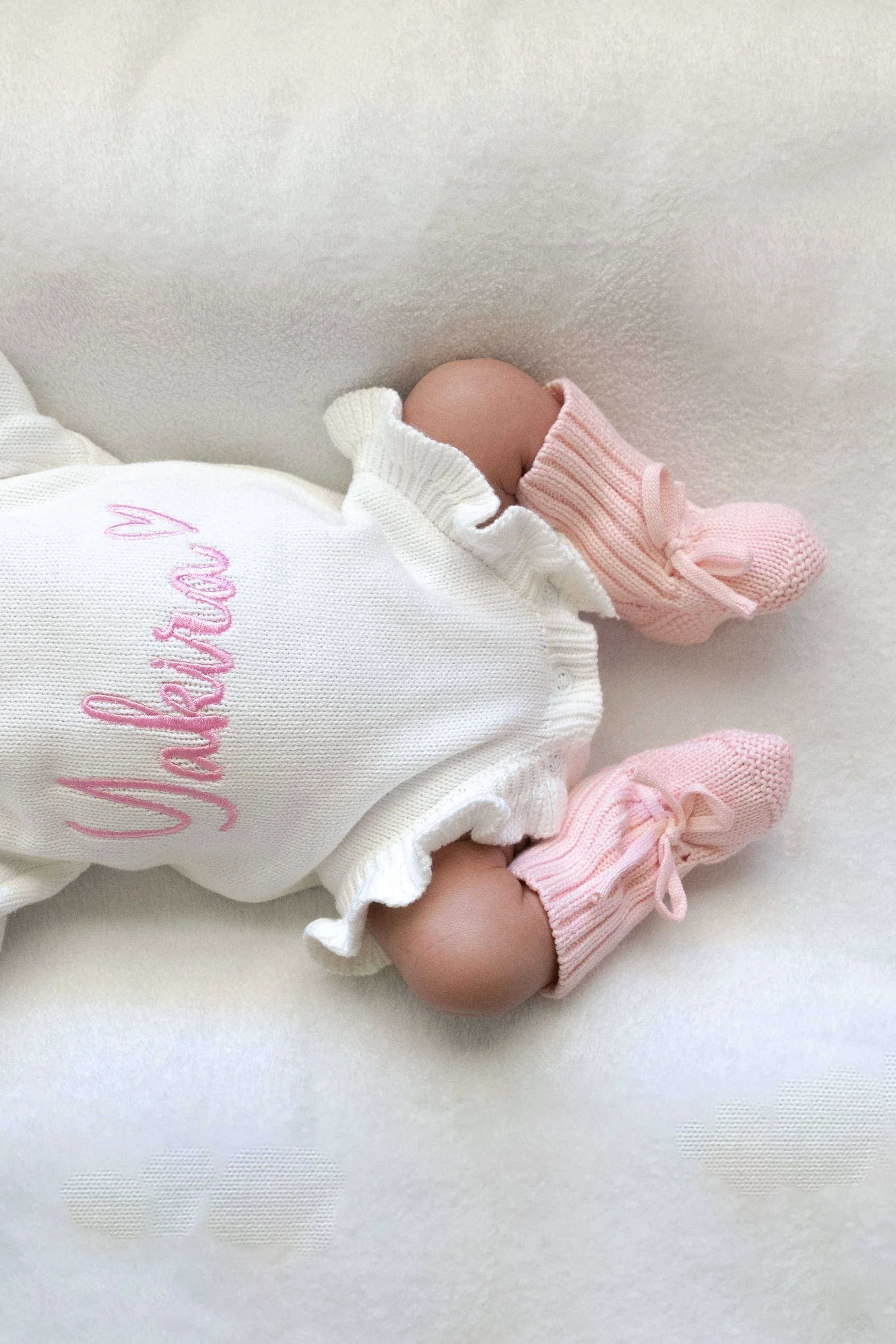 Close-up of a baby lying on a white surface, wearing pink knitted socks and a white outfit with pink embroidery that reads 'Valentina'.
