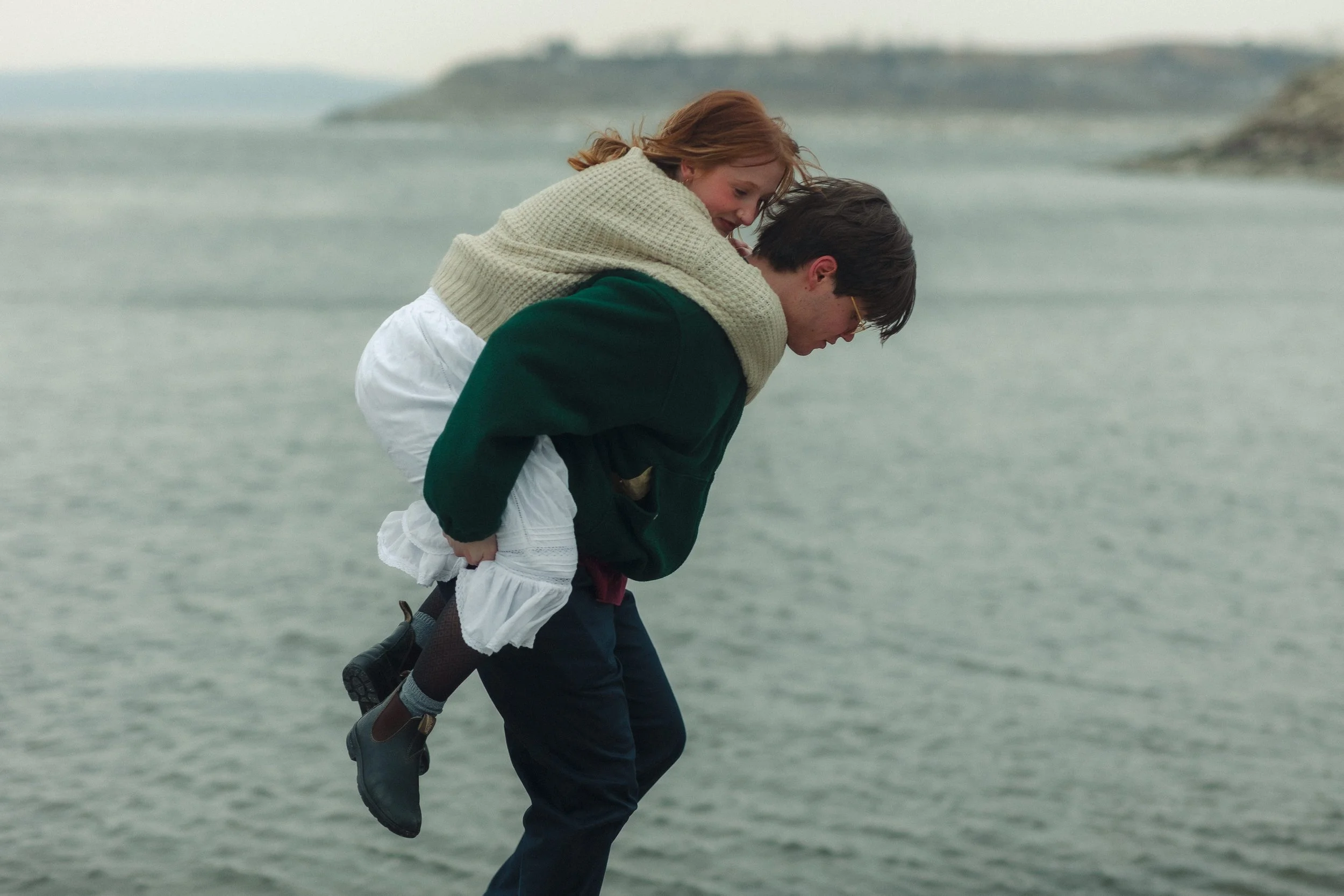 A young woman with red hair seen riding on the back of a young man near a body of water, with a landscape in the background.
