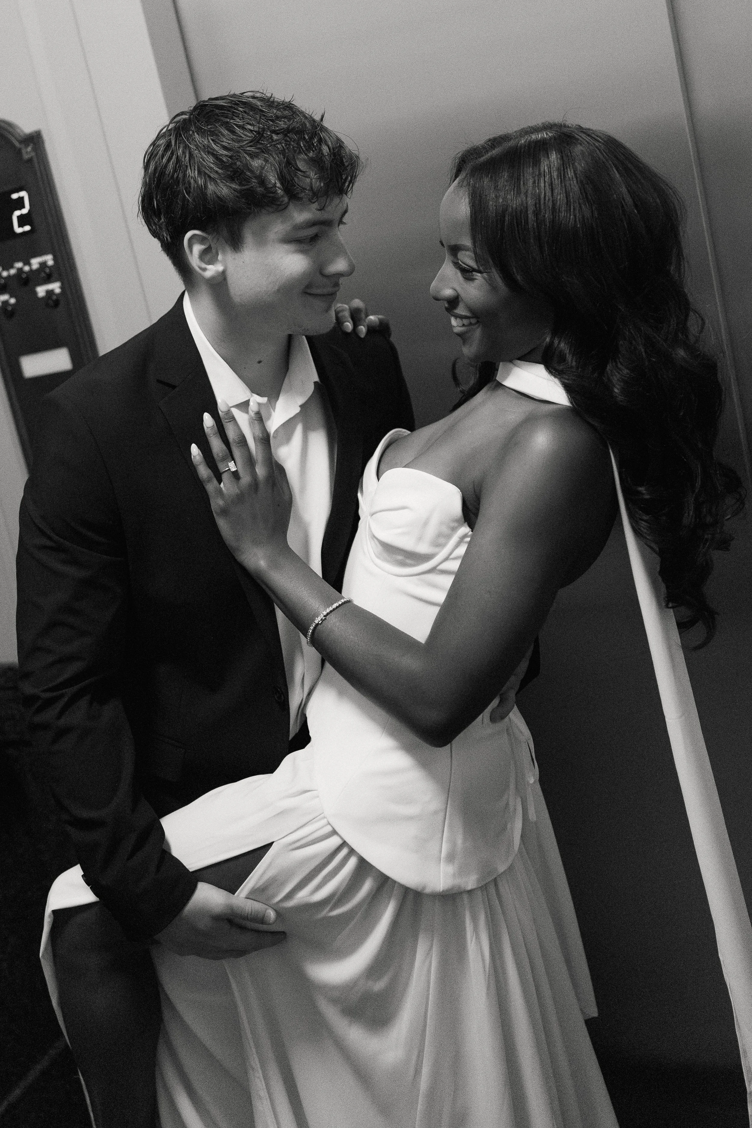 A black-and-white photo of a smiling man in a suit and a woman in a dress, standing close together in an elevator, facing each other, with the woman's hand on the man's chest and the man's hand on the woman's waist.