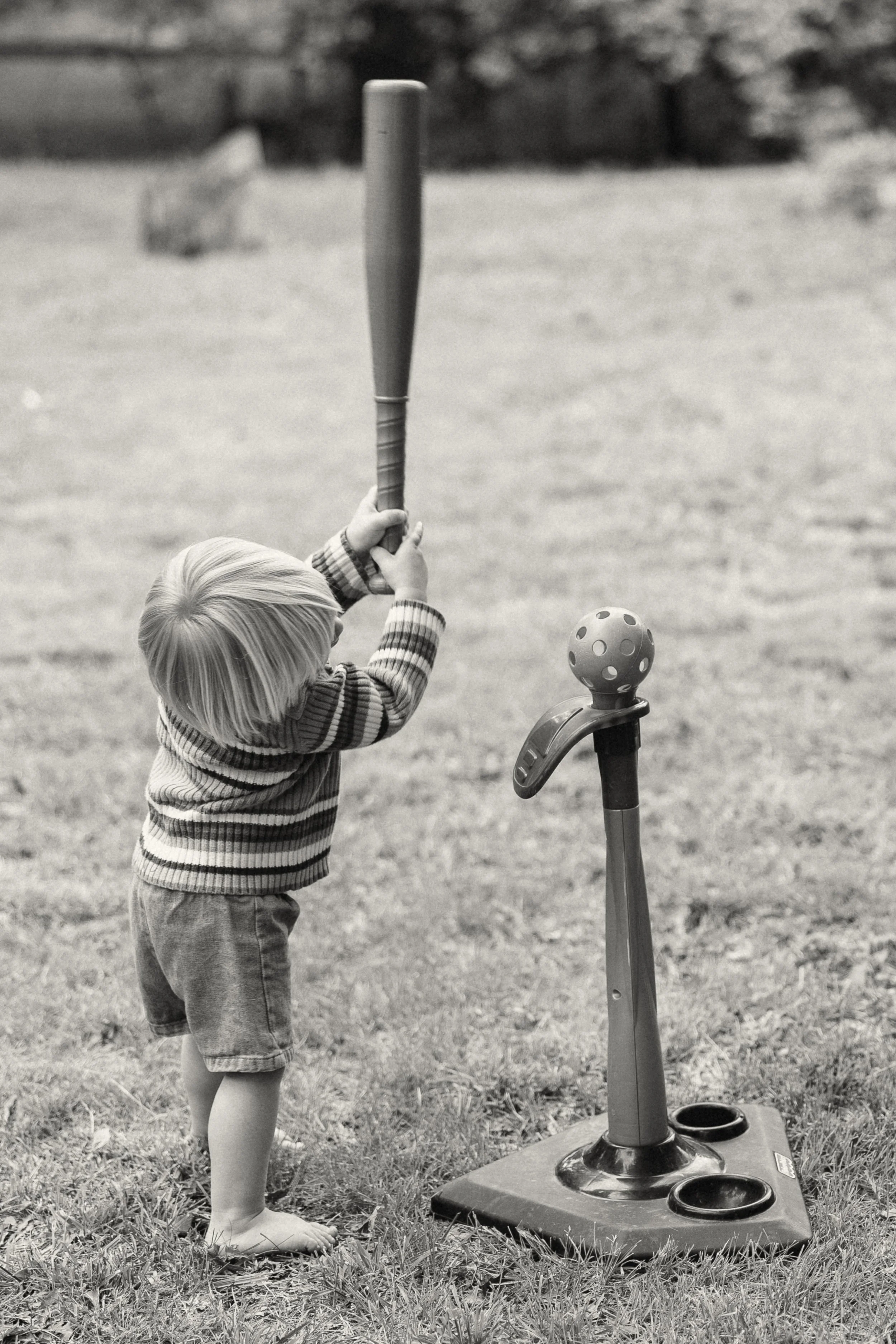 A young child with light hair, wearing a striped sweater and shorts, barefoot in a grassy outdoor area, holding a baseball bat and preparing to hit a ball from a pitching machine.