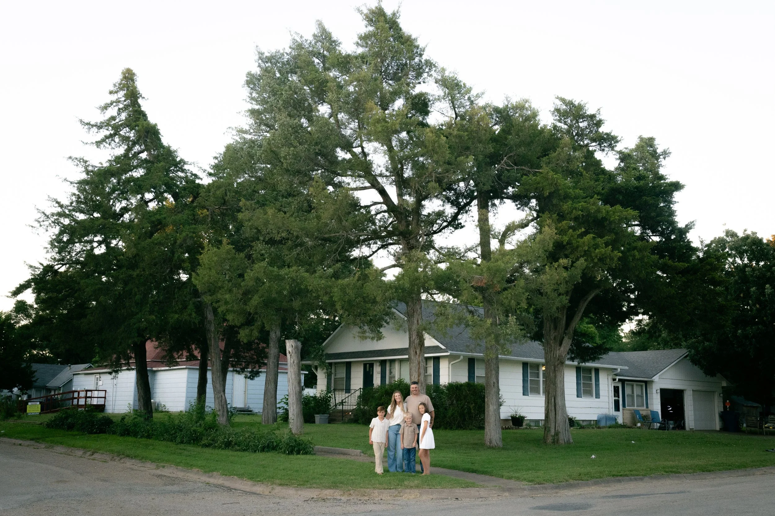 A family of five standing on a sidewalk in front of a white house with trees in the yard.