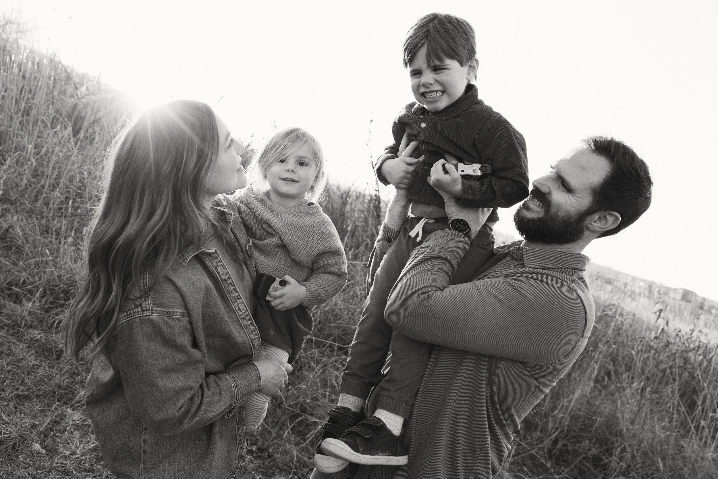 A family of four outdoors, with the father lifting a young boy while a woman and girl look on, all smiling or making funny faces, in a grassy area with a bright sky.