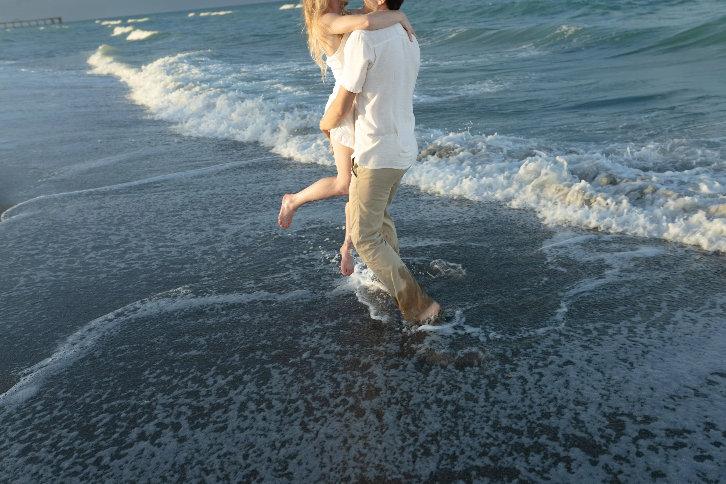 A person in khaki pants and a white shirt holding a child in a white dress, both barefoot, playing in the shallow water at the beach with small waves.