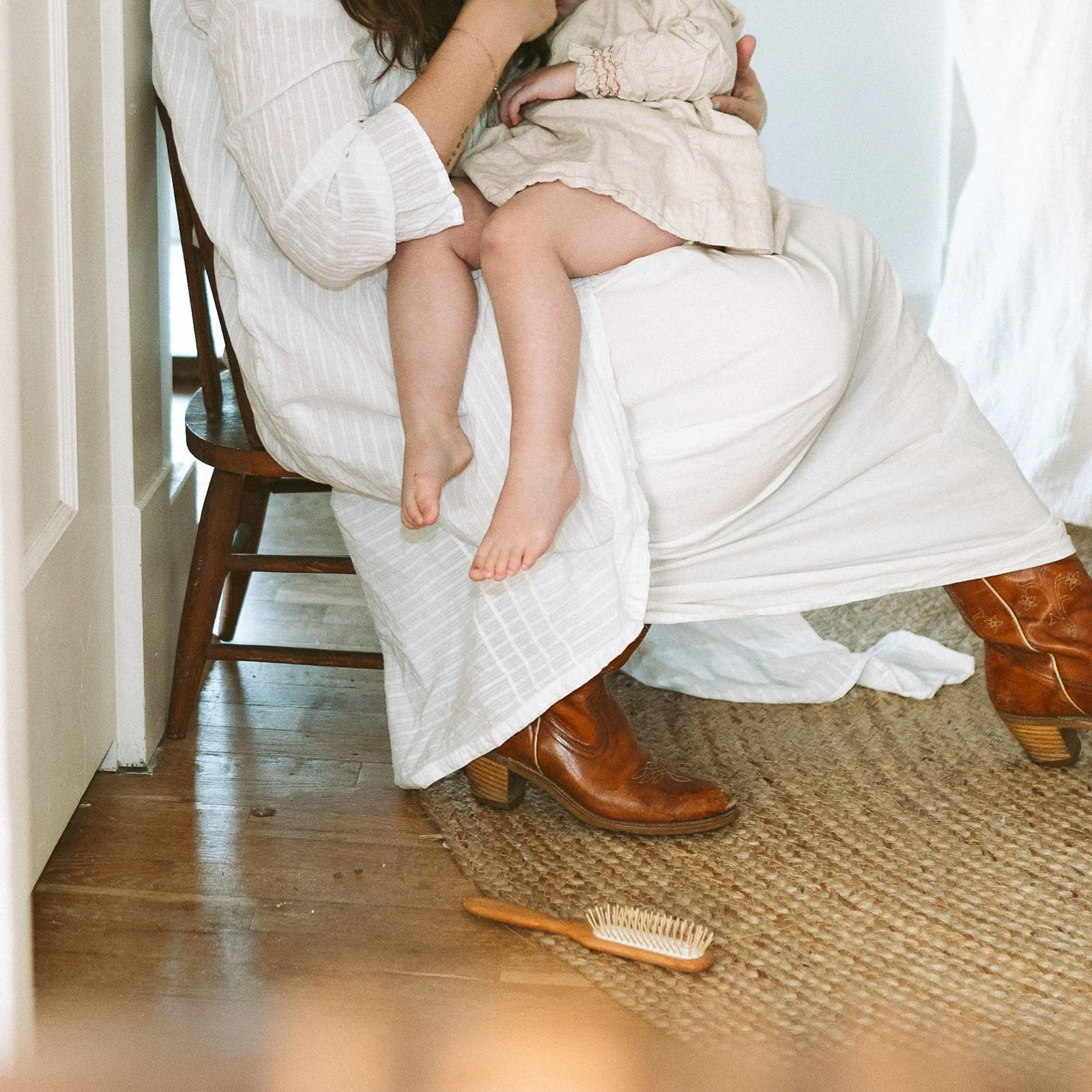 A woman and a child sitting on a wooden chair, with the woman holding the child. The woman is wearing white and brown cowboy boots. There is a hairbrush on the wooden floor nearby and a woven rug.