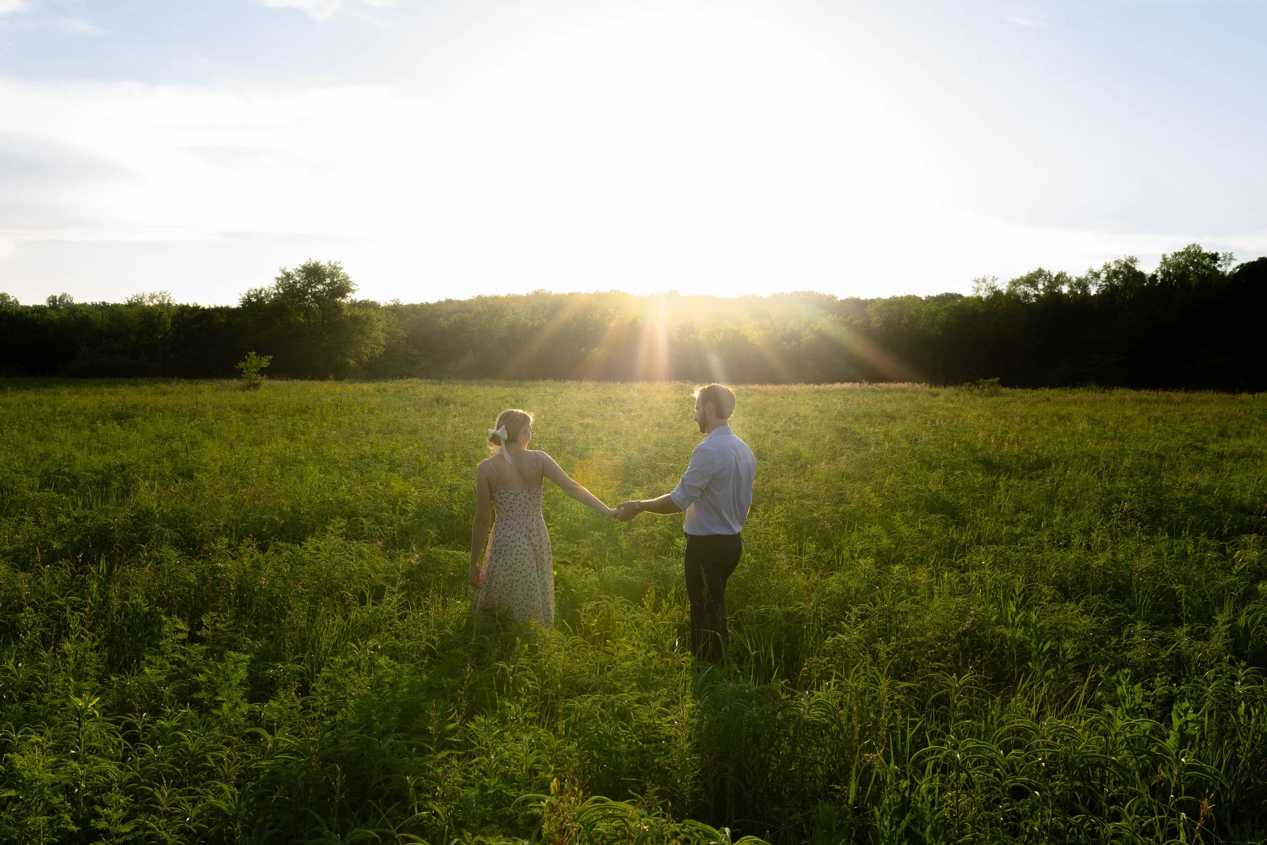 A couple holding hands and walking through a green field at sunset with the sun shining brightly above the horizon.