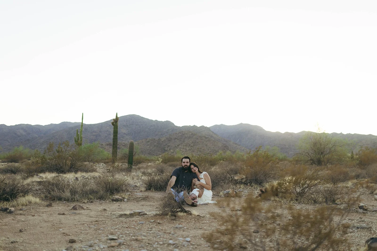 A couple sitting on the desert ground, surrounded by bushes and cacti, with mountains in the background and a bright sky.