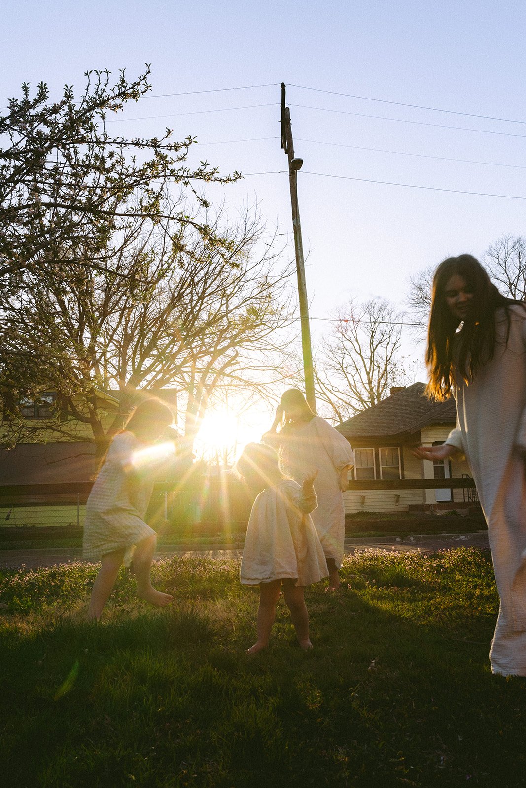 Four young girls playing outdoors on a grassy area during sunset, with trees and houses in the background.