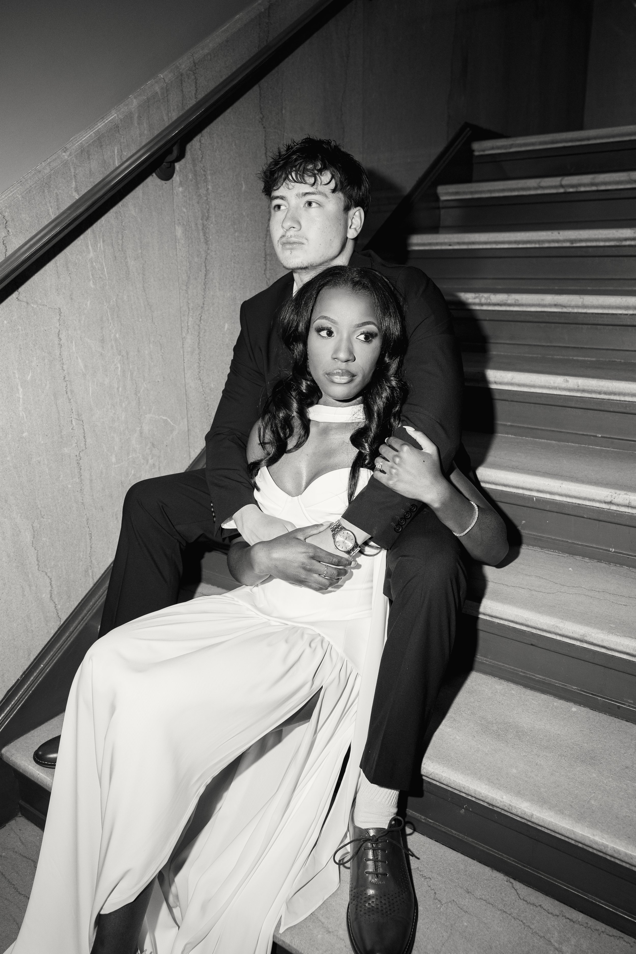 Two young adults, a man and a woman, sitting on stairs indoors. The man has dark, tousled hair and light skin, dressed in a dark jacket. The woman has long, wavy dark hair, glasses, and darker skin, wearing a white dress. The woman is sitting on the 