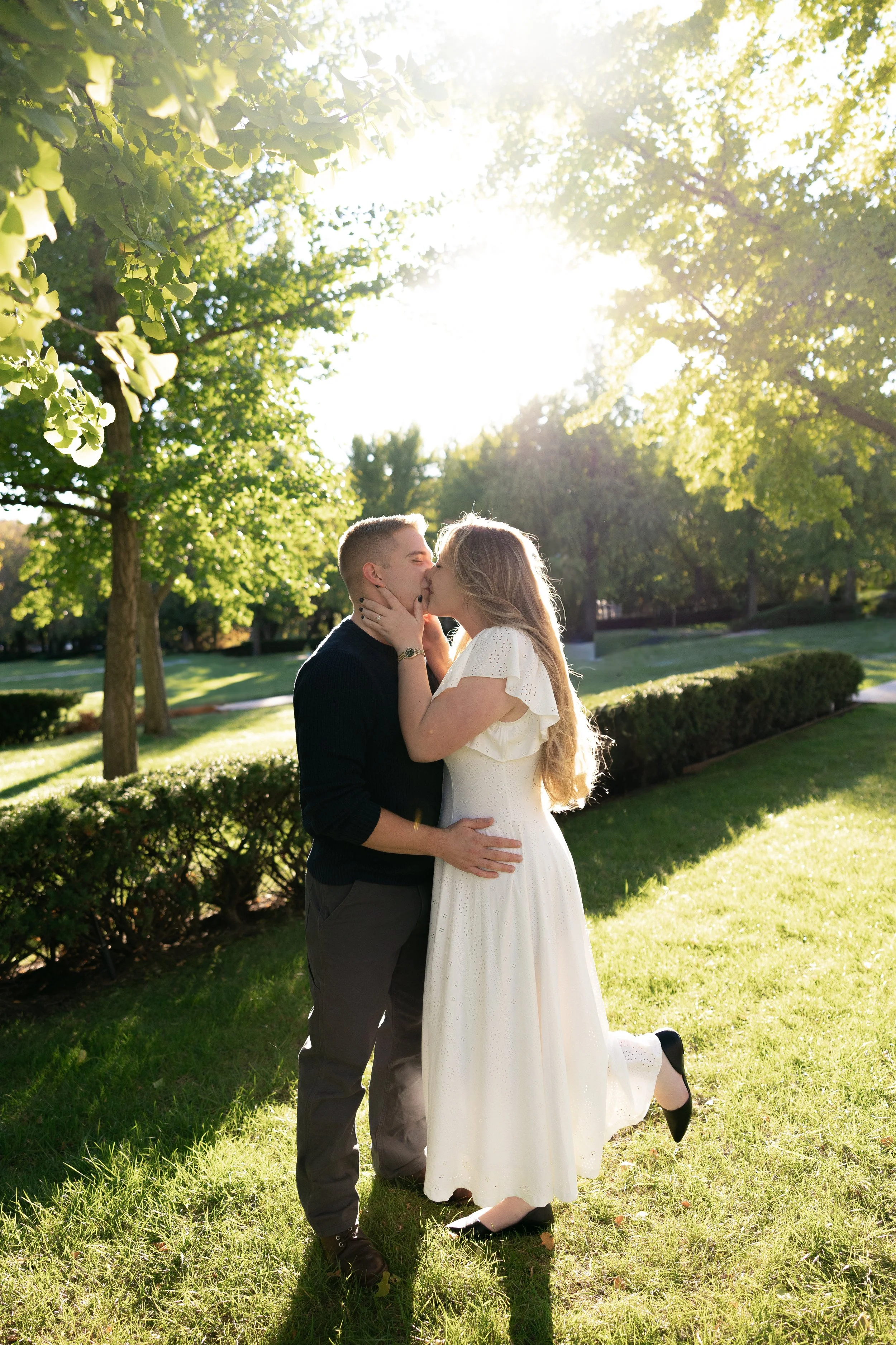 A couple kissing in a park on a sunny day, with trees and grass in the background.