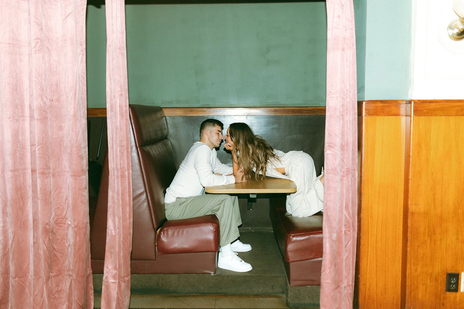 A young couple is sitting face to face in a booth of a restaurant, leaning in close and smiling, with pink curtains framing the scene.