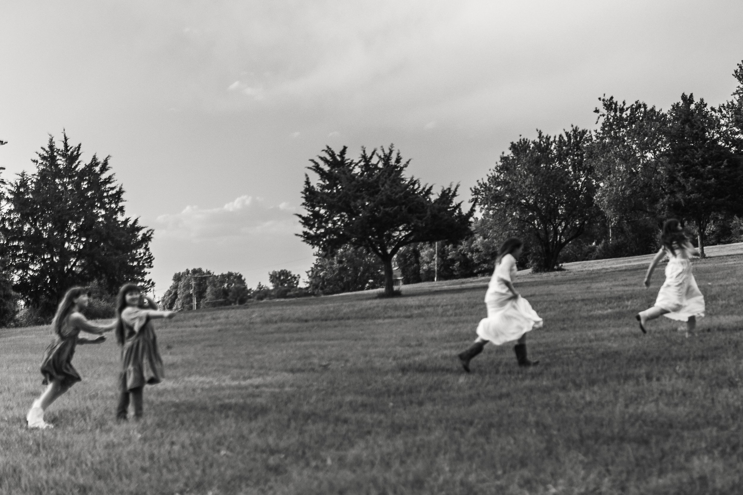 Black and white photo of five girls running and playing on a grassy hillside, with trees and clouds in the background.