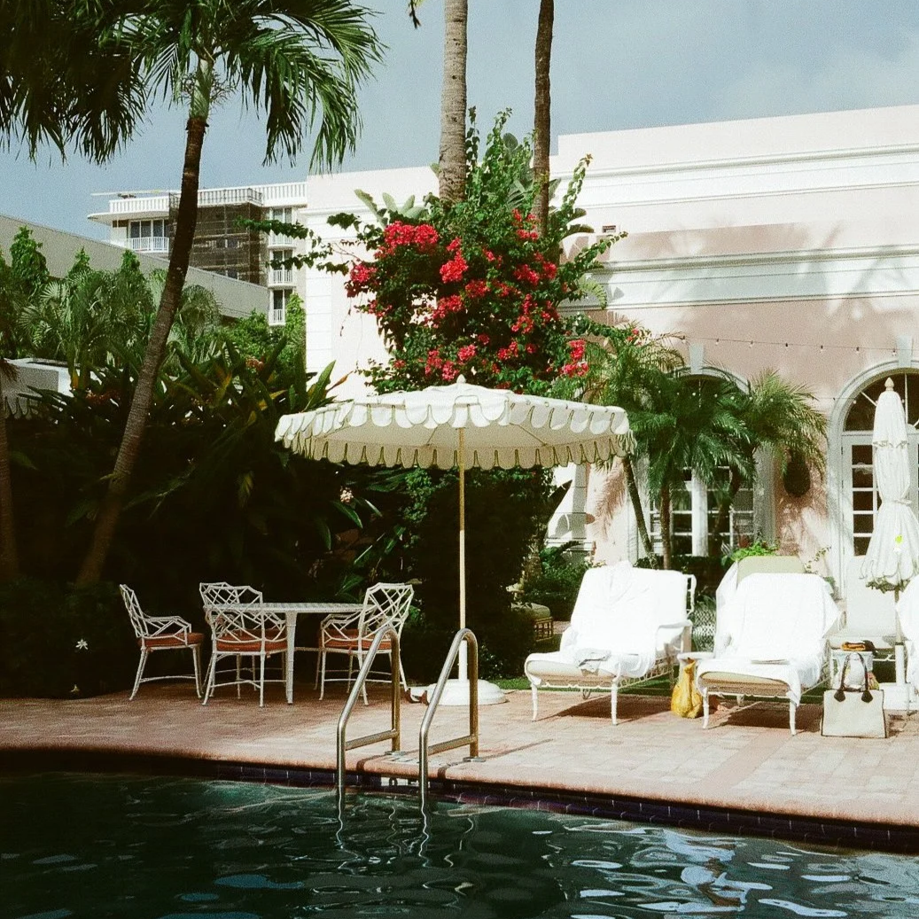 Poolside with white lounge chairs, an umbrella, tropical plants, and a pink building in the background.