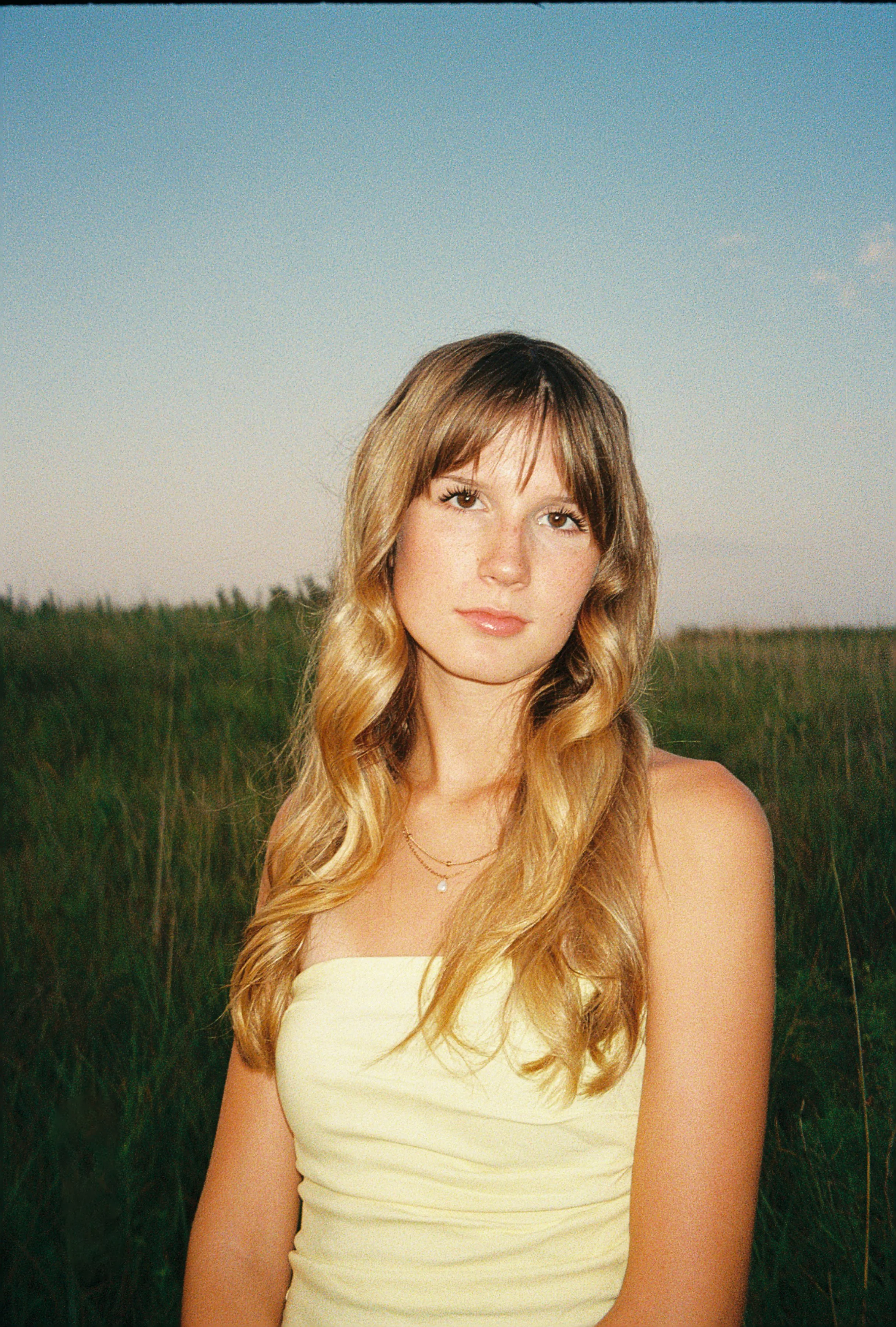 A young woman with long, wavy blonde hair wearing a strapless yellow top and layered necklaces standing outdoors in a grassy field during sunset.