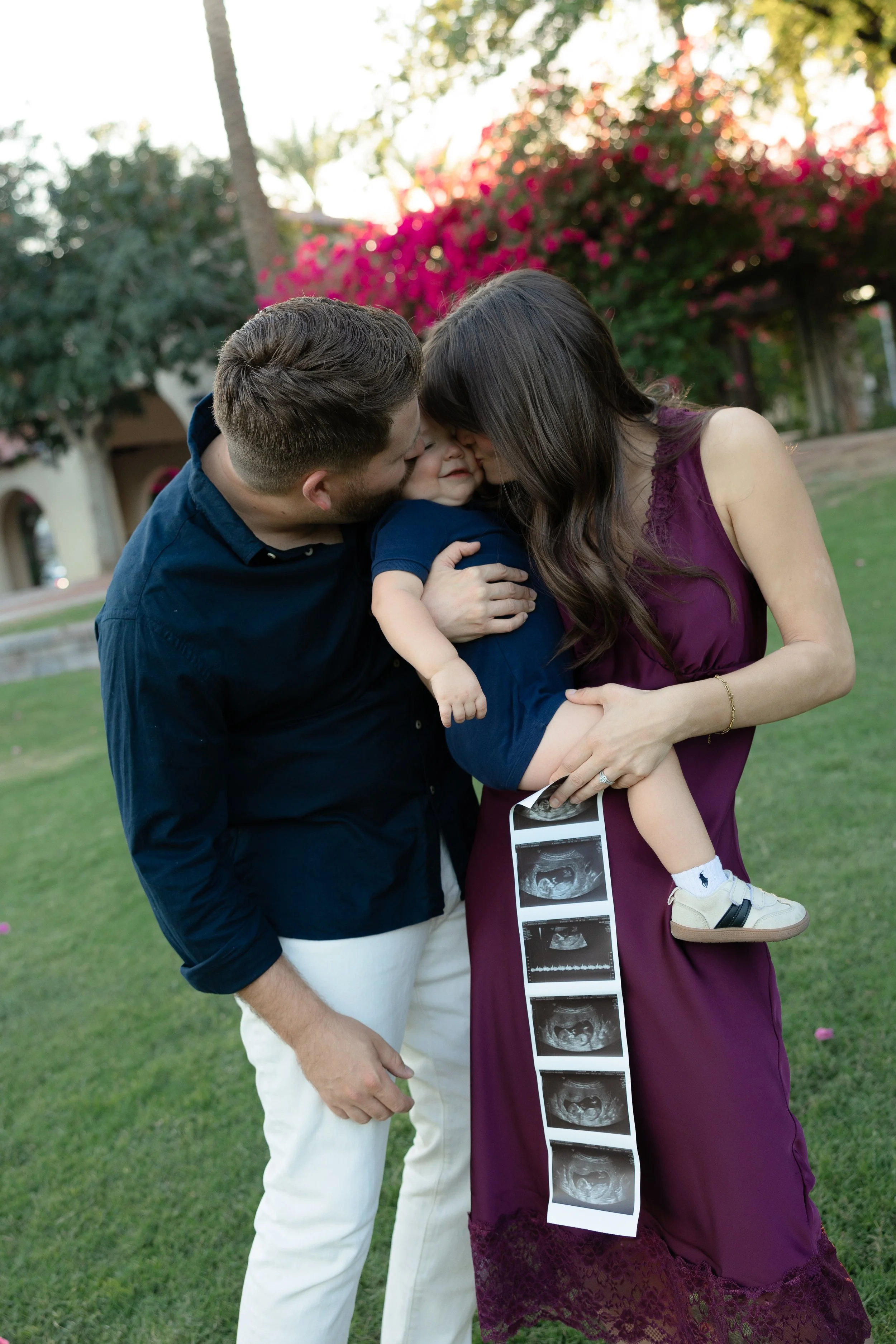 A family of three, a man, a woman, and a small child, sharing a moment of affection outdoors. The woman is holding ultrasound images.