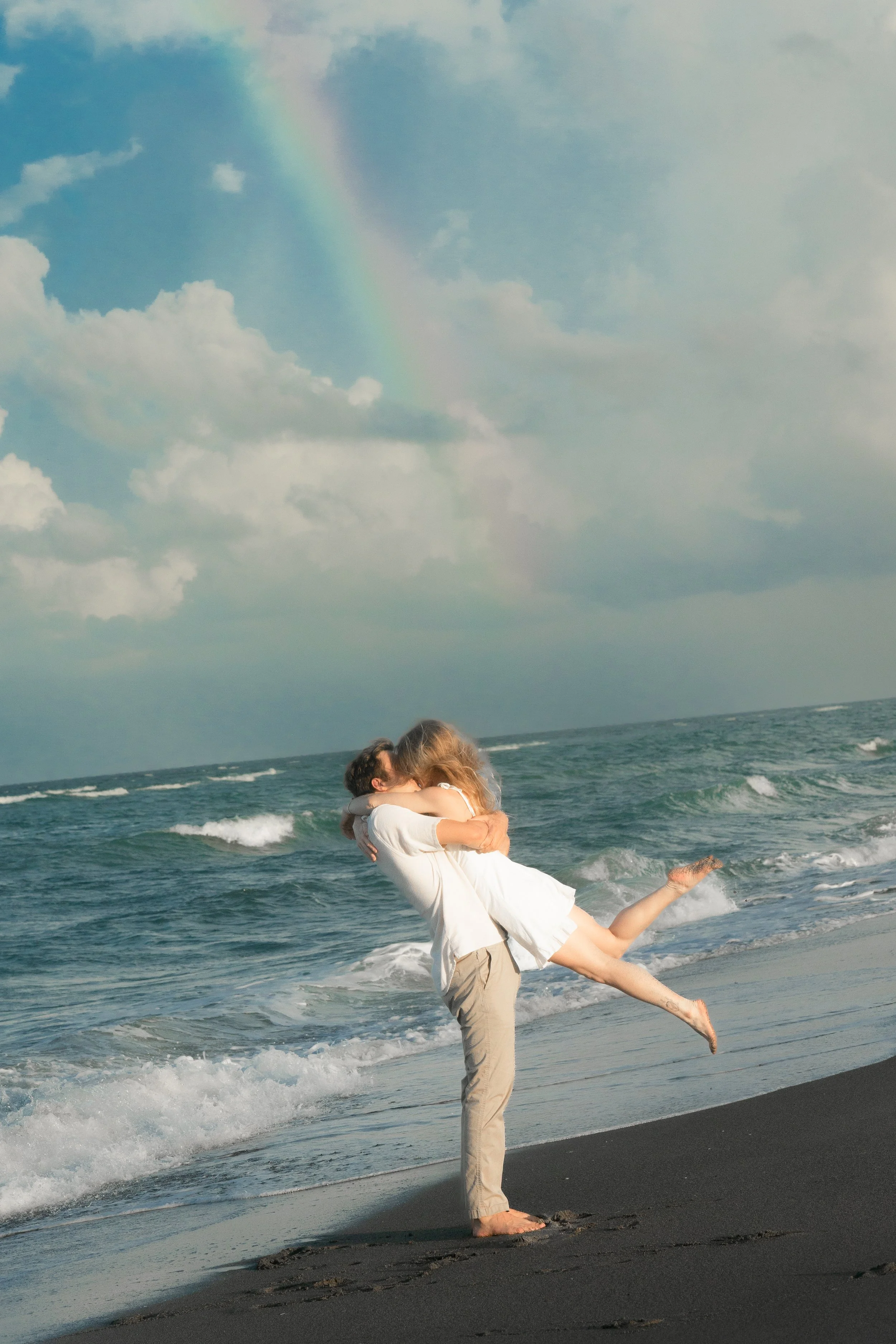 A couple on the beach, with the man lifting and hugging the woman, and a rainbow in the sky.