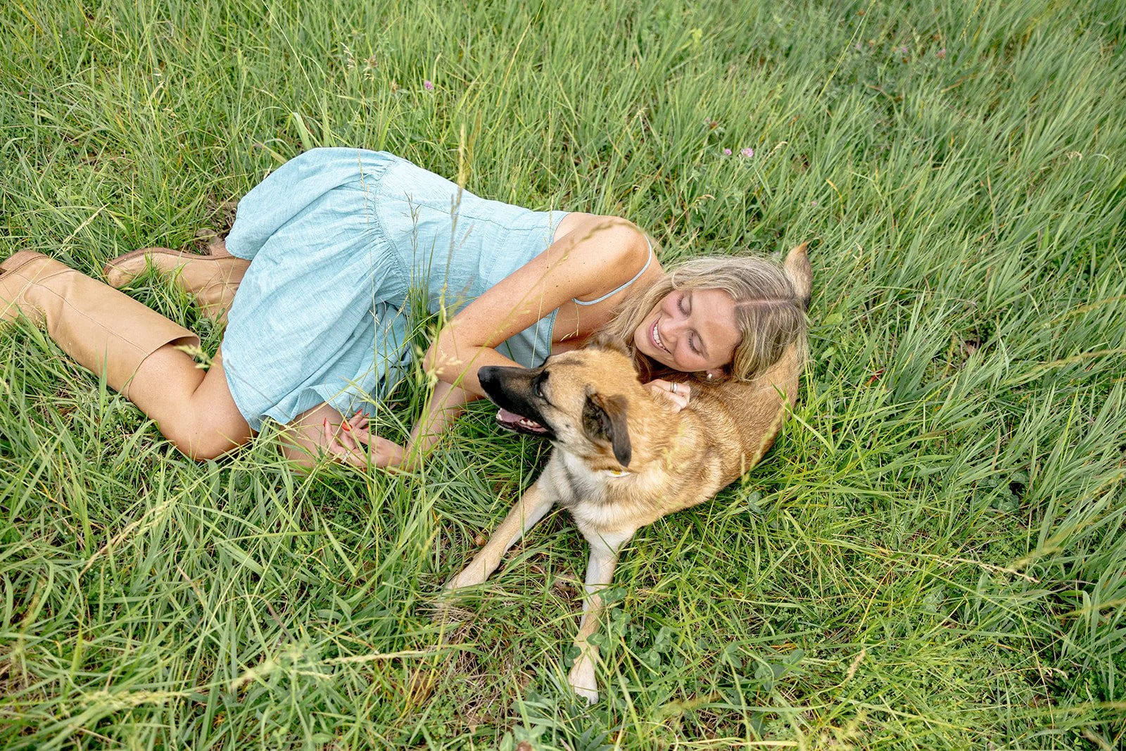 A woman lying on her side in tall green grass smiling and cuddling with a brown and black dog.