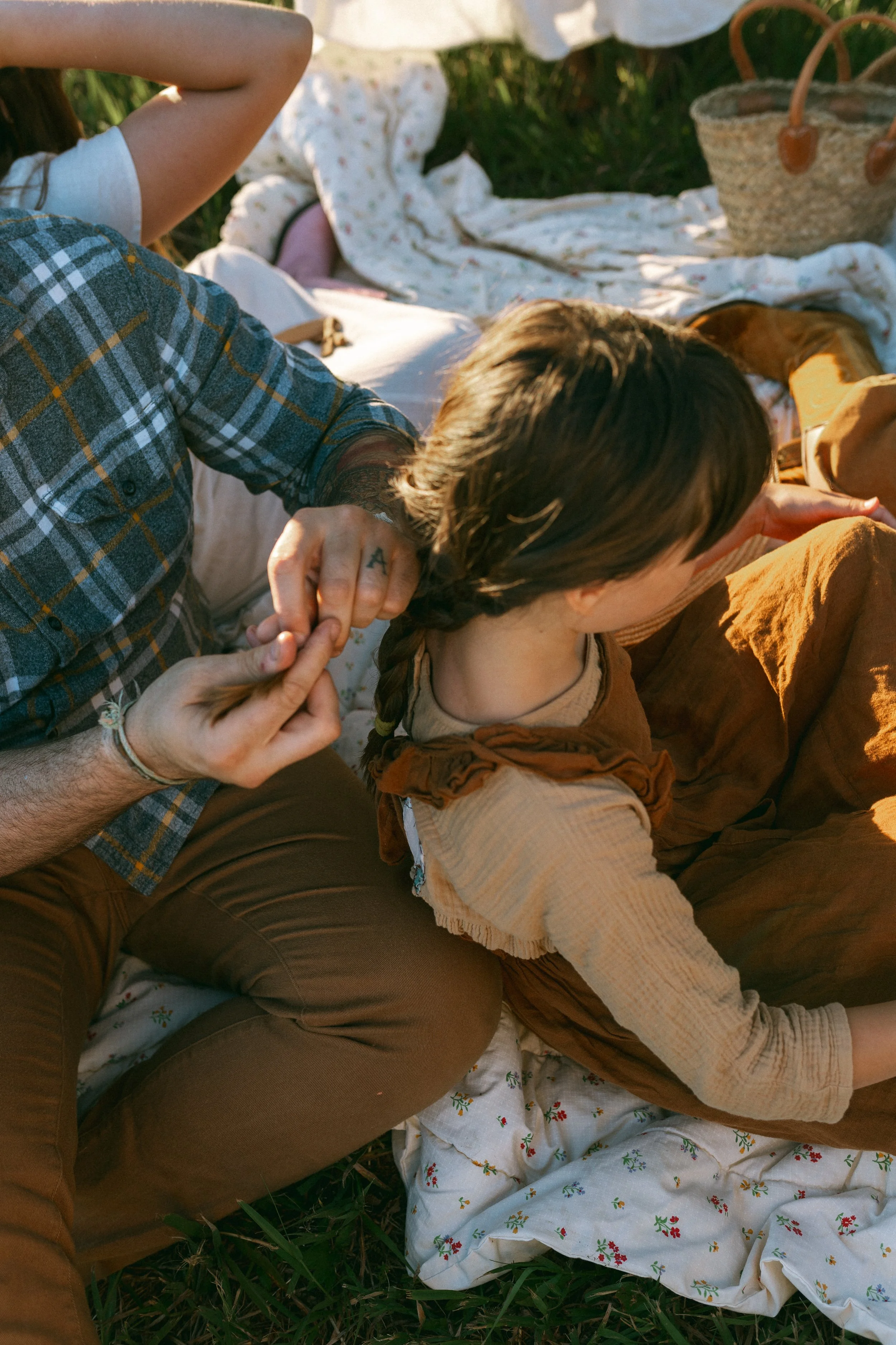 A person with tattoos and a plaid shirt is holding a young girl with braids and long sleeve shirt, sitting on a picnic blanket outdoors on grass, with picnic baskets and fabric blankets in the background.