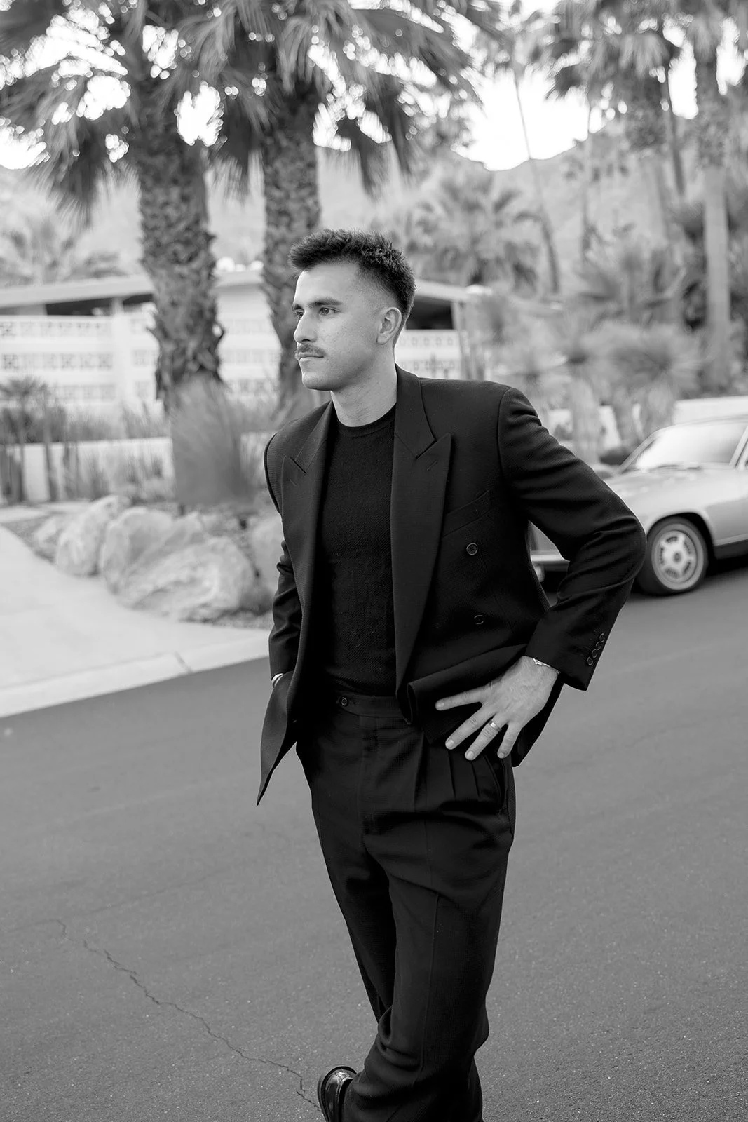 A man in a black suit and black shirt standing outdoors on a street with palm trees and cars in the background, captured in black-and-white.