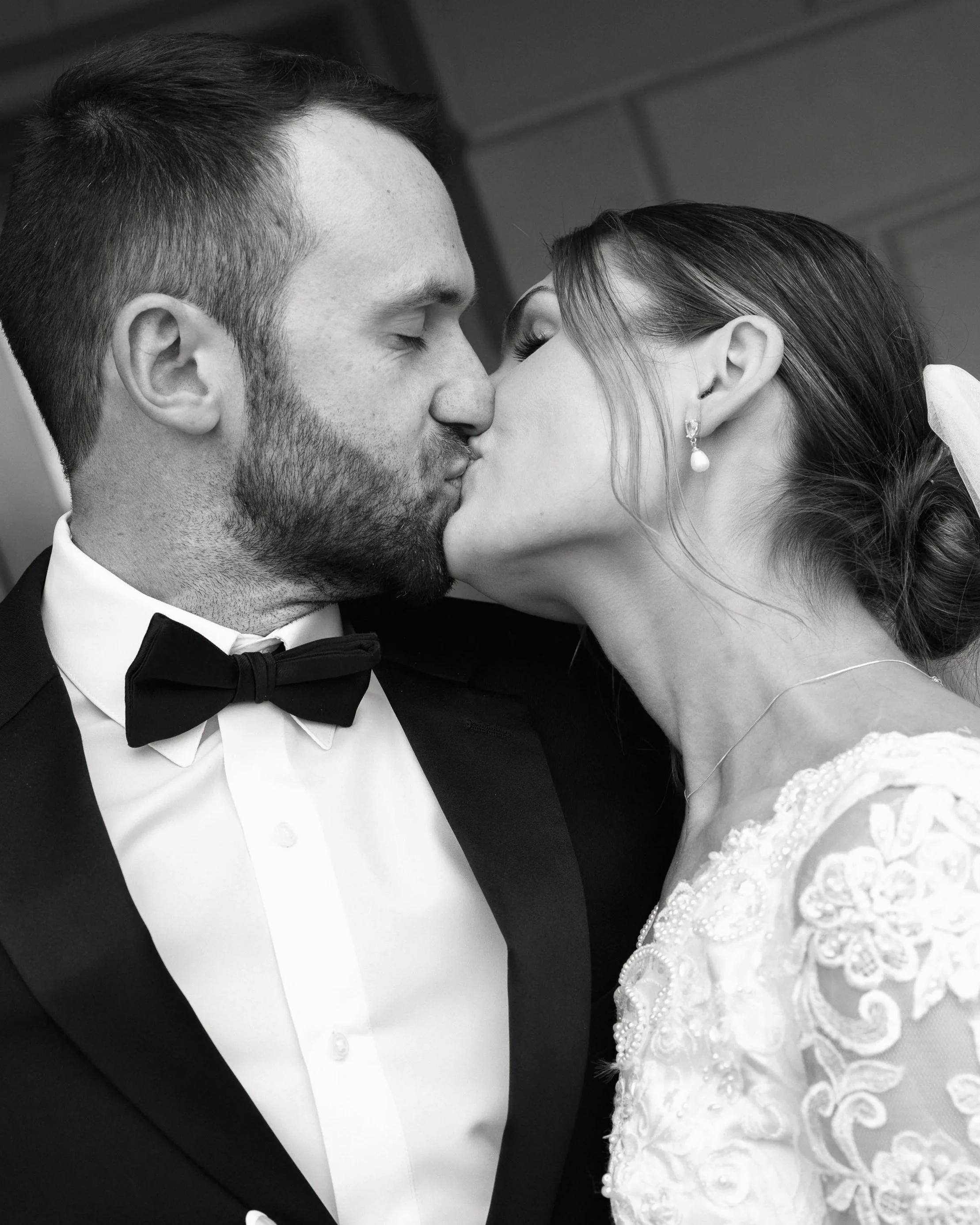 A black and white photo of a bride and groom sharing a kiss. The groom is wearing a tuxedo with a bow tie and the bride is wearing a wedding dress with intricate lace and pearls, with pearl earrings.