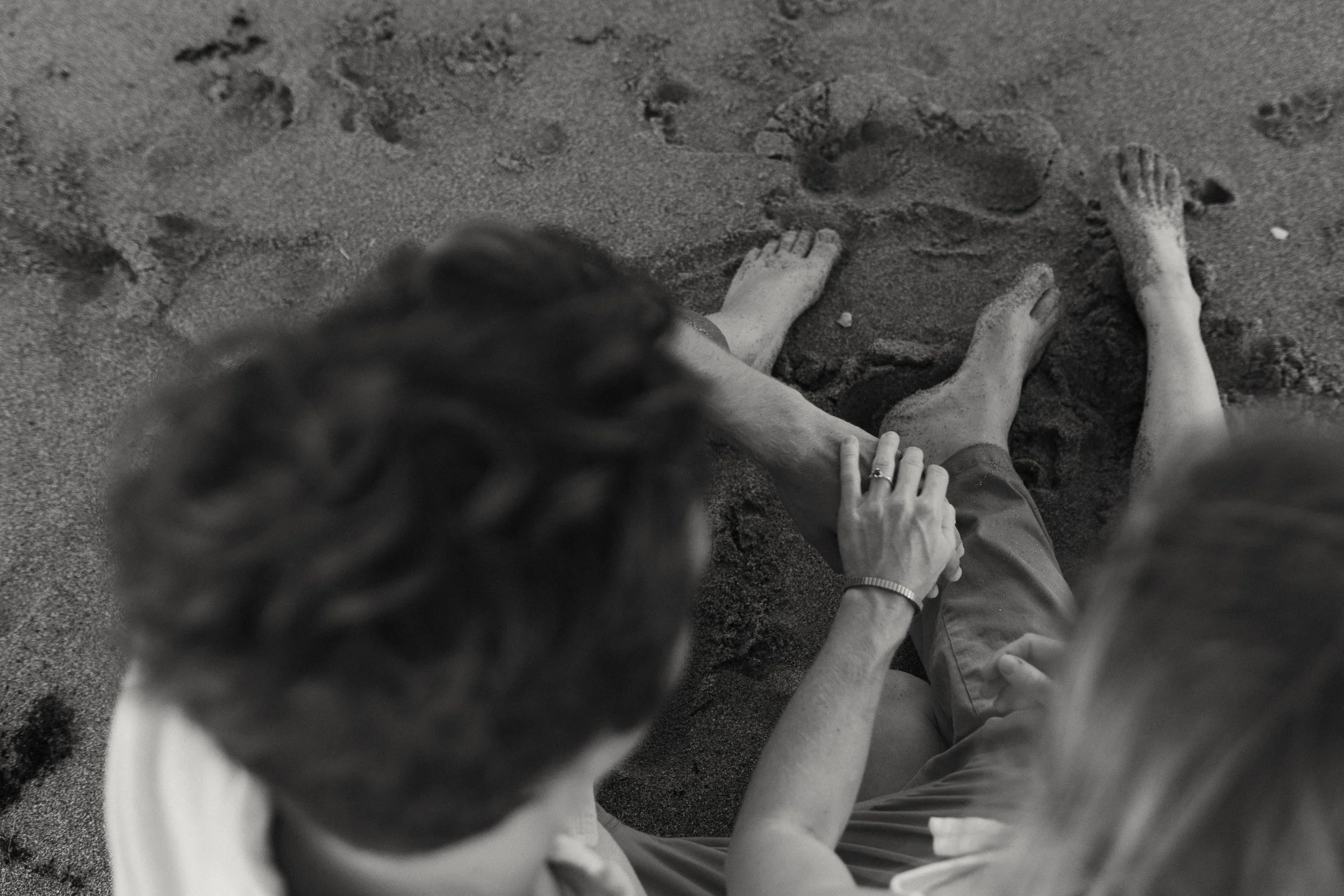 A black and white photo of two people sitting on a sandy beach, holding hands, with their feet in the sand and visible footprints nearby.