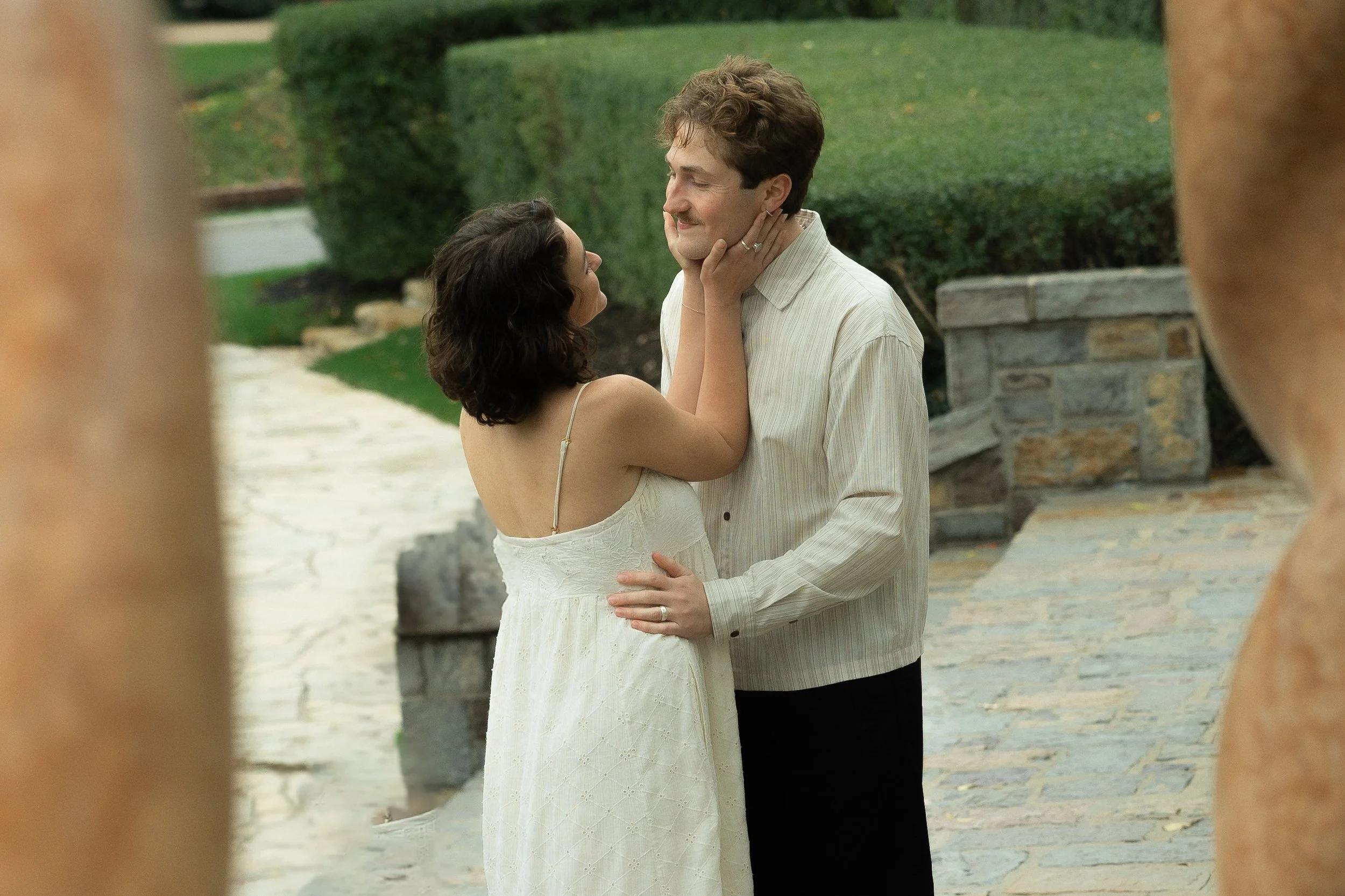 A couple sharing a tender moment outdoors, with the woman gently holding the man's face and the man smiling warmly, framed by two blurred stone pillars.