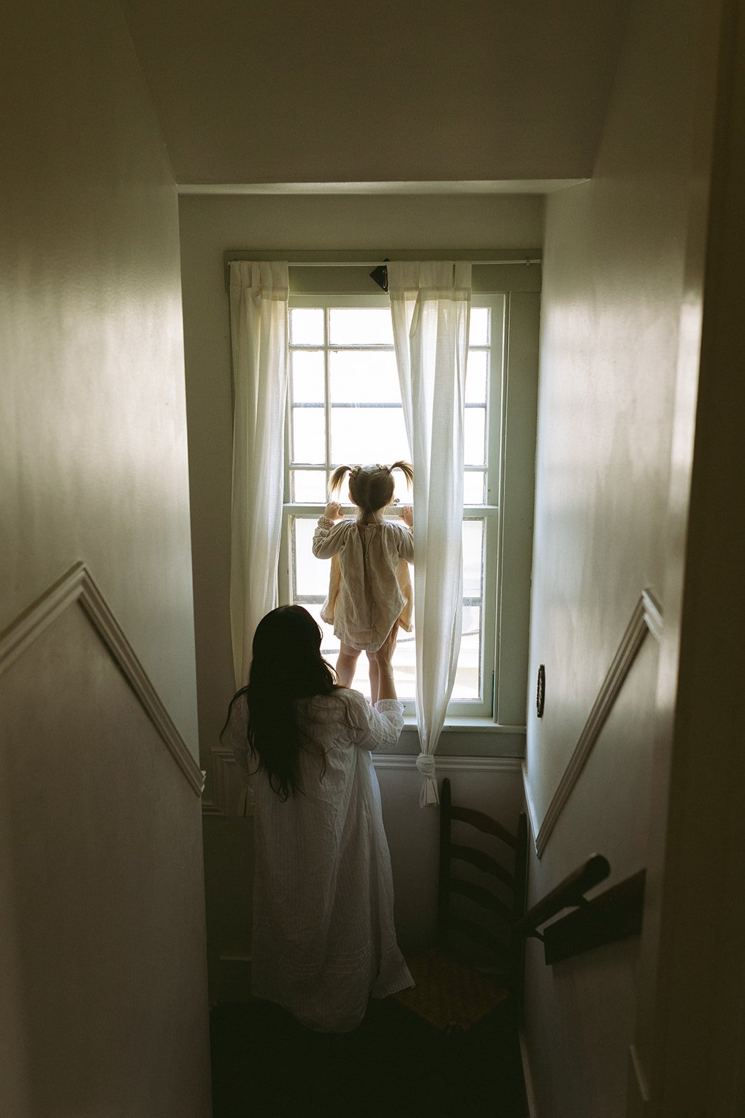 A woman with long dark hair in a white dress standing behind a young girl with a ponytail, also in a white dress, looking out a large window with white curtains in a cozy, softly lit room.