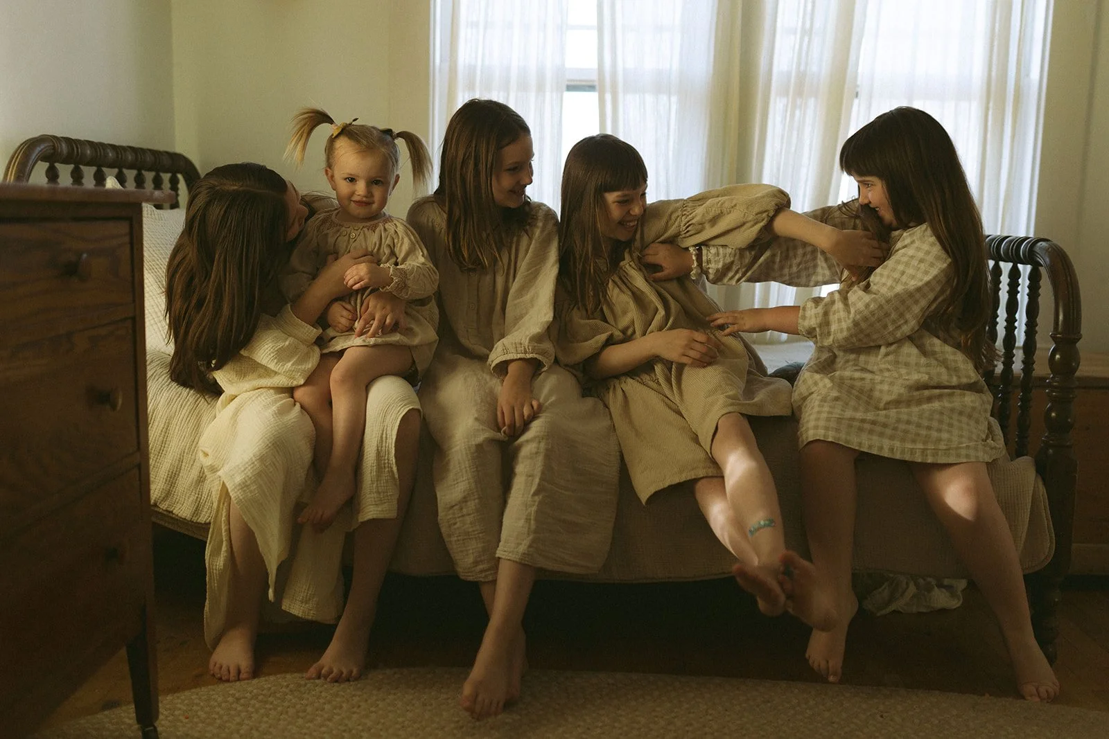 Seven young girls in beige and checkered nightgowns playing and tickling each other on a bed in a warmly lit bedroom.