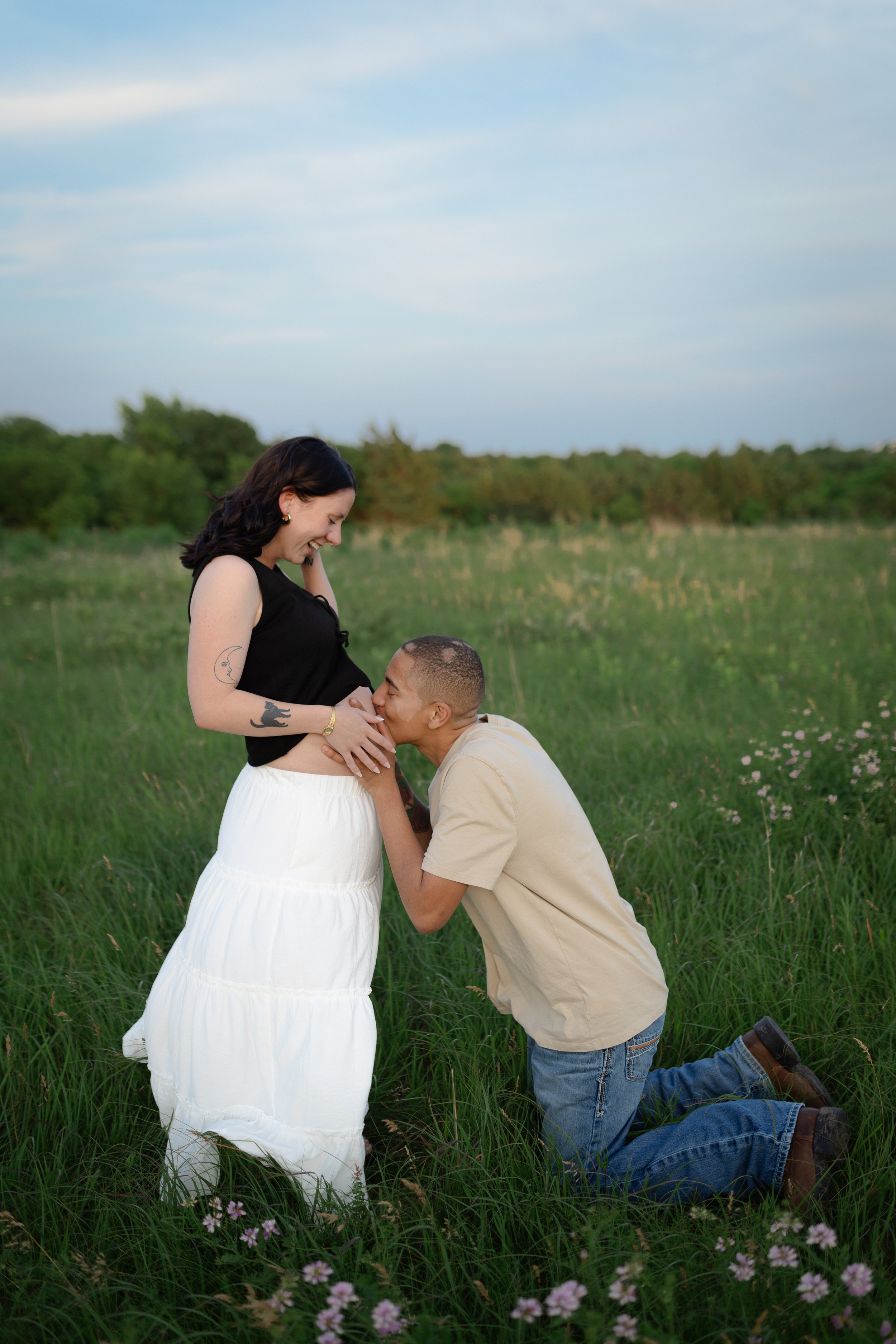 A man proposes to a woman in a grassy field, kneeling with a ring and the woman smiling joyfully.