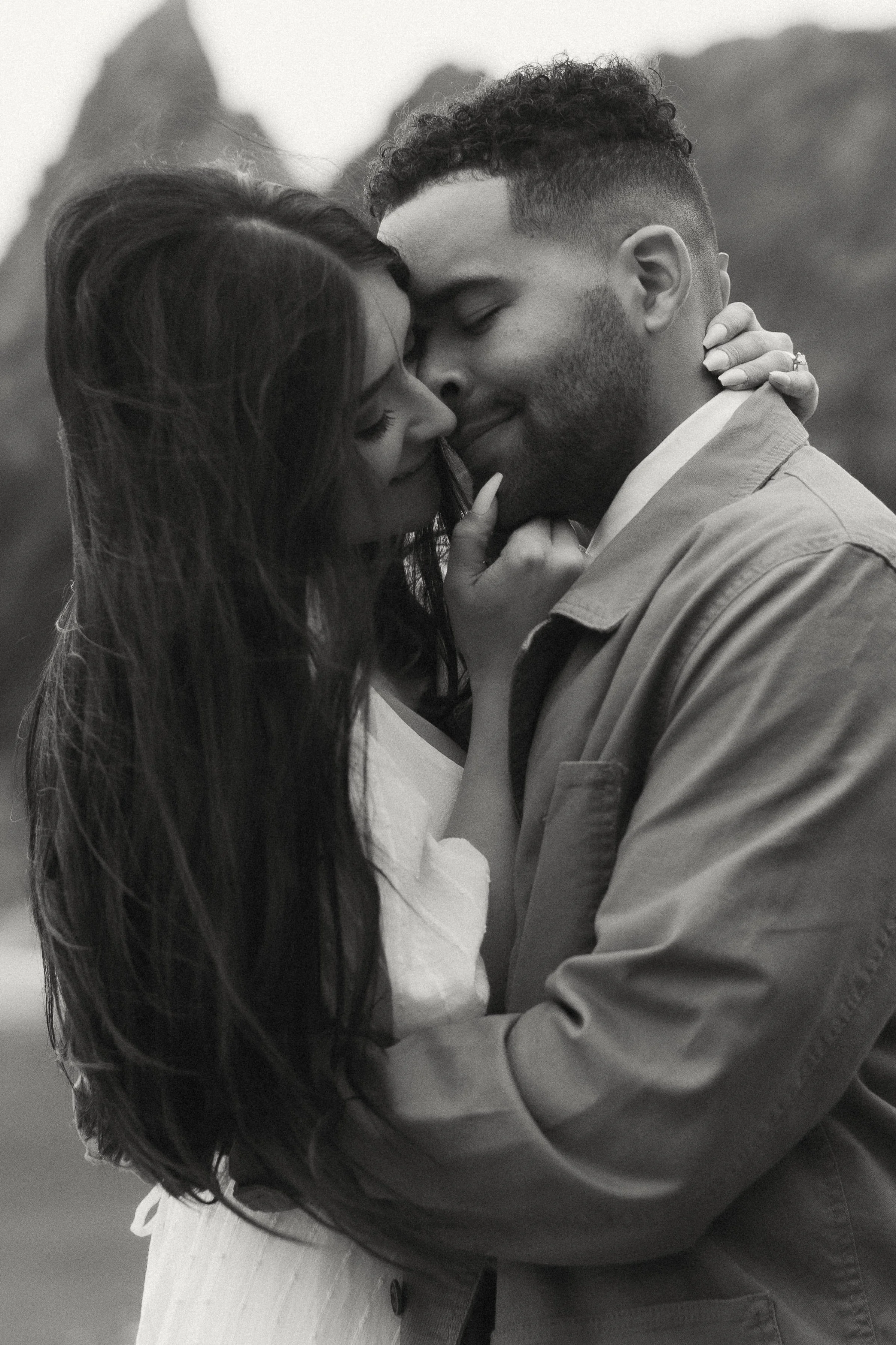 A black-and-white photo of a couple embracing tenderly, with their foreheads touching, in an outdoor setting with mountains in the background.