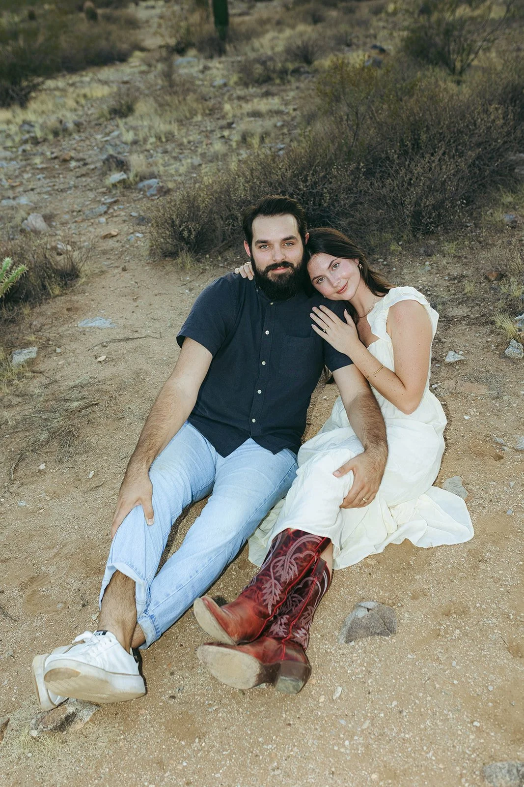 A couple sitting on a dirt trail in a desert landscape, embracing and smiling at the camera.