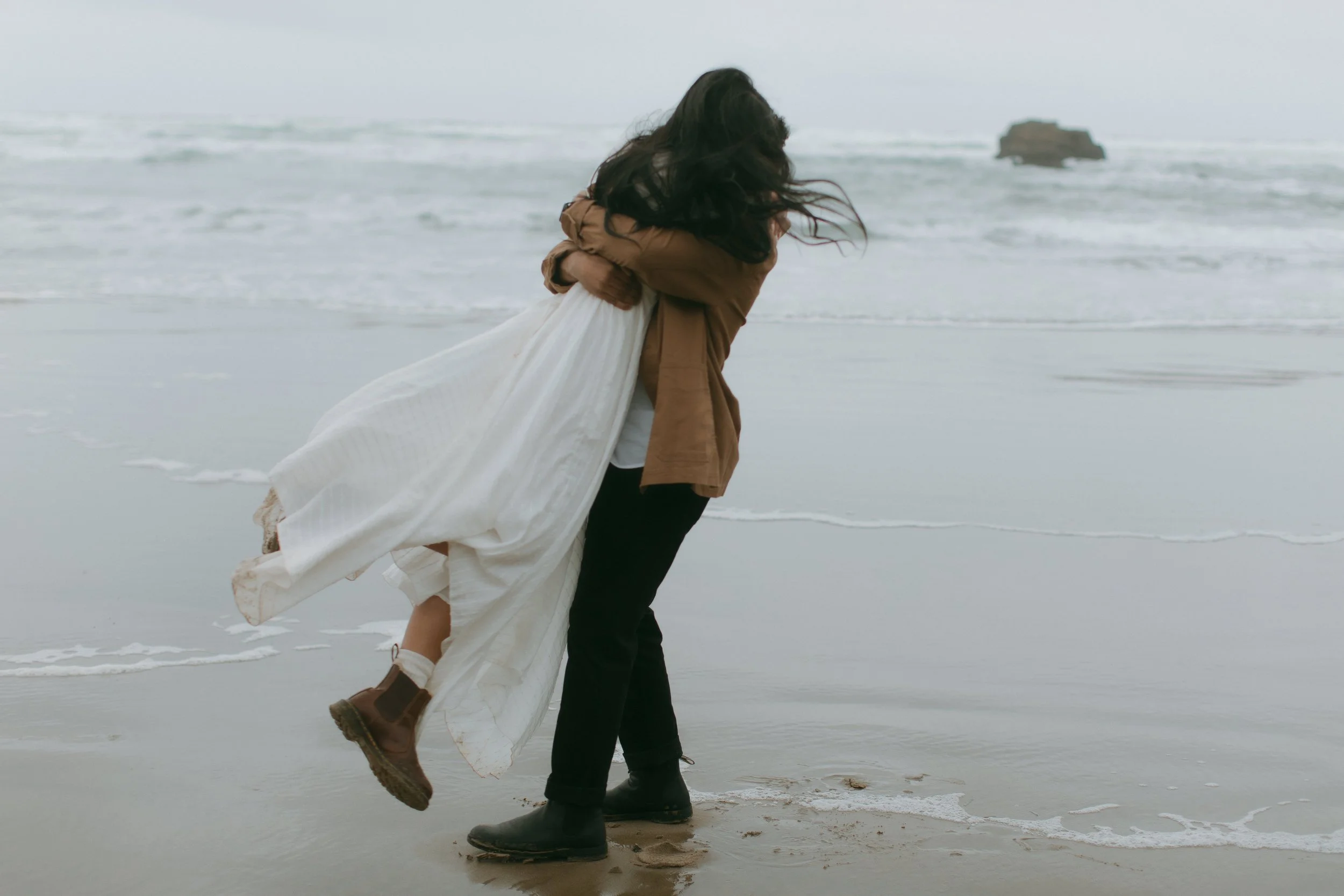 A person is holding a woman in a white dress on a beach with ocean waves in the background.