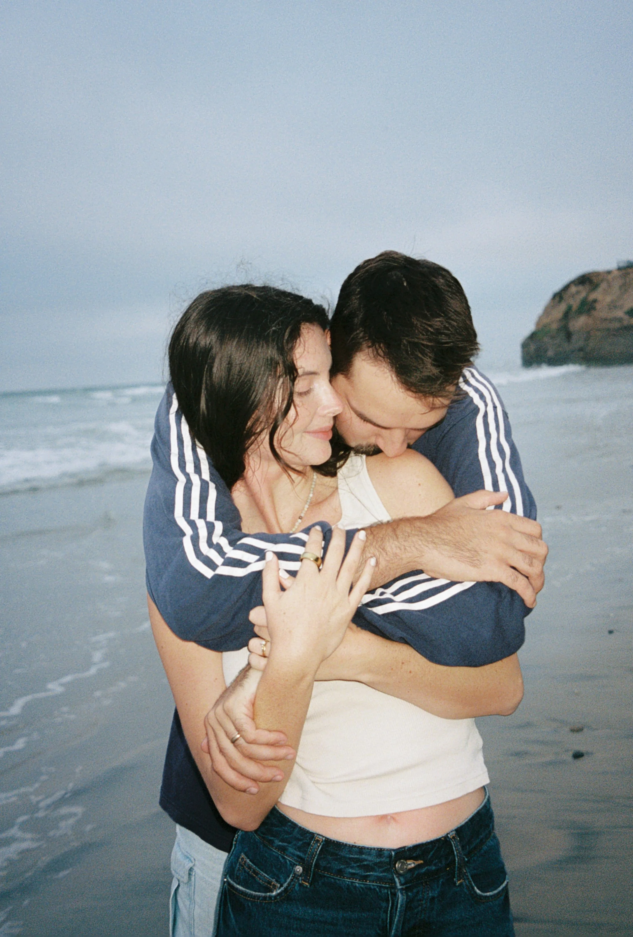 A couple embracing on a beach, with the man hugging the woman from behind and the woman smiling with her eyes closed.