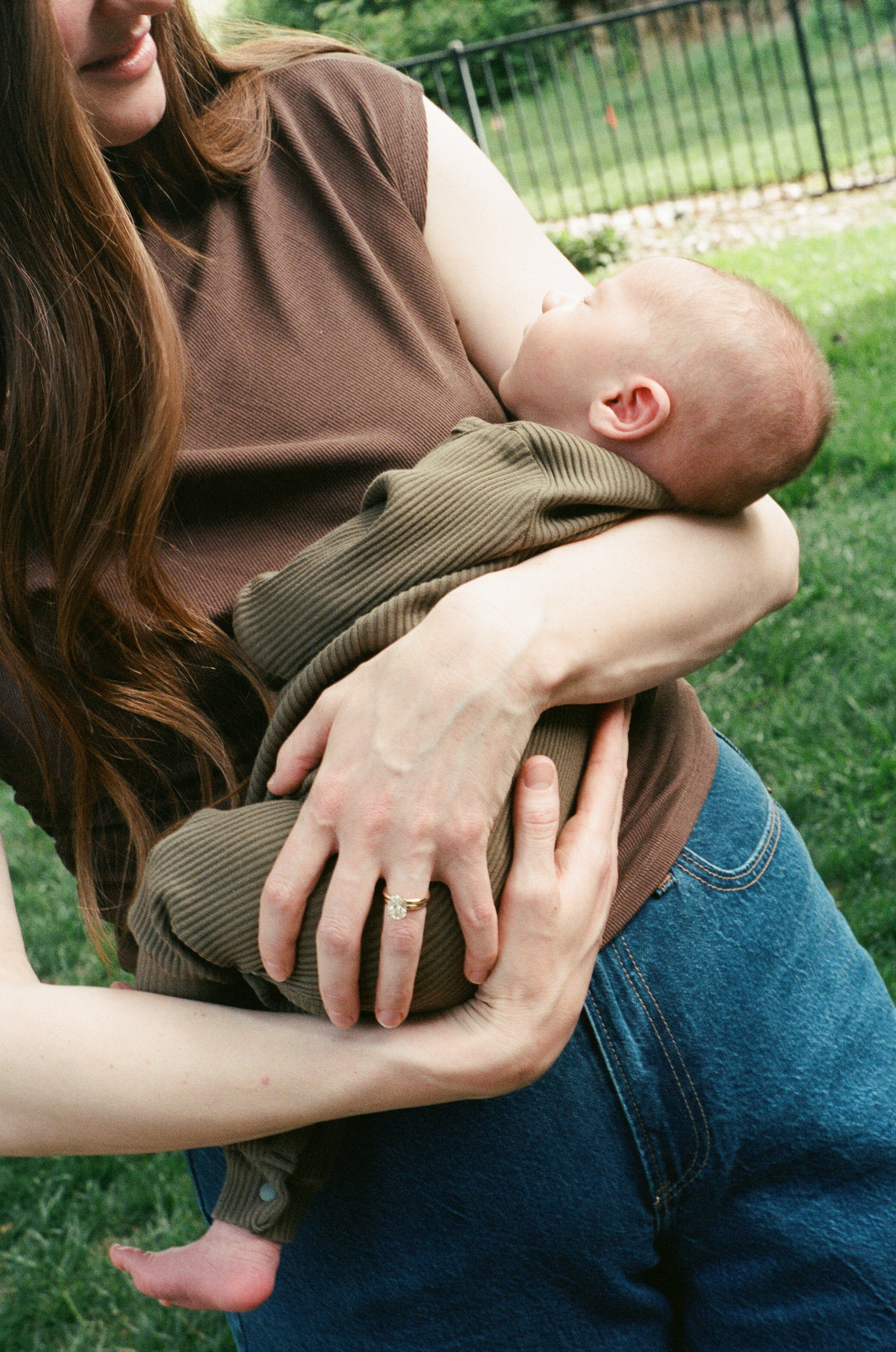 A woman holding a baby outdoors on a grassy area near a metal fence.