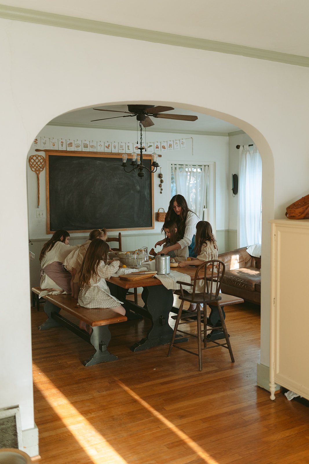 A woman and six children gathered around a dining table in a cozy, sunlit kitchen, preparing food.