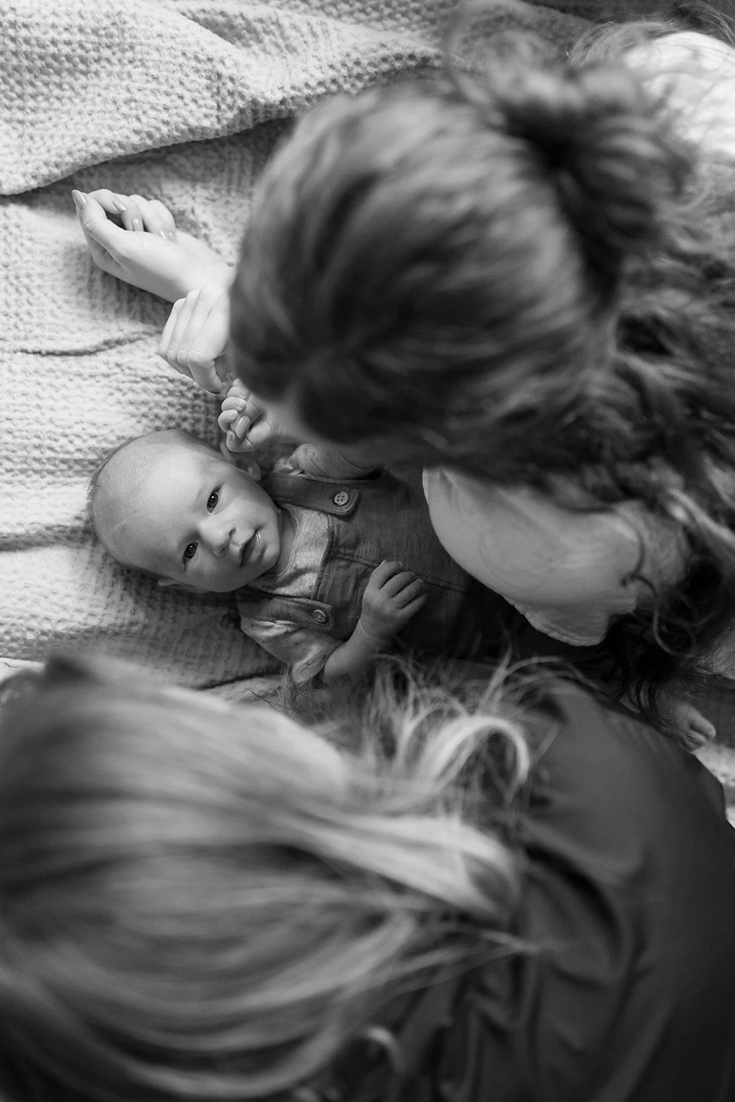 A black-and-white photo shows a woman lying on her back holding a baby's hand, with the baby looking up at her. The woman has long, wavy hair, and the baby is lying on a textured blanket.
