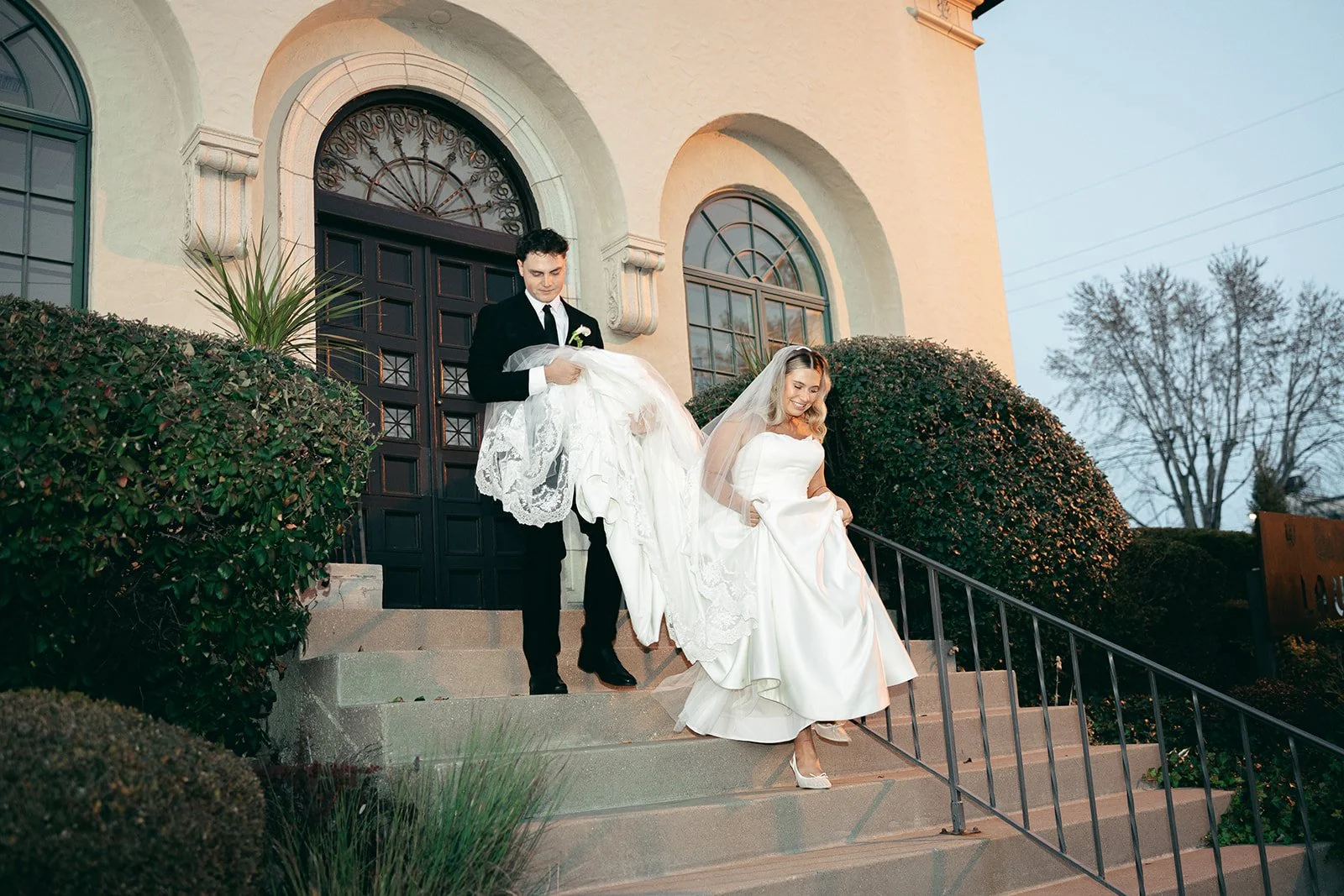 A bride in a white wedding dress and veil is descending outdoor stairs with a groom in a black suit and tie carrying her dress. They are outside a church with arched windows and bushes.