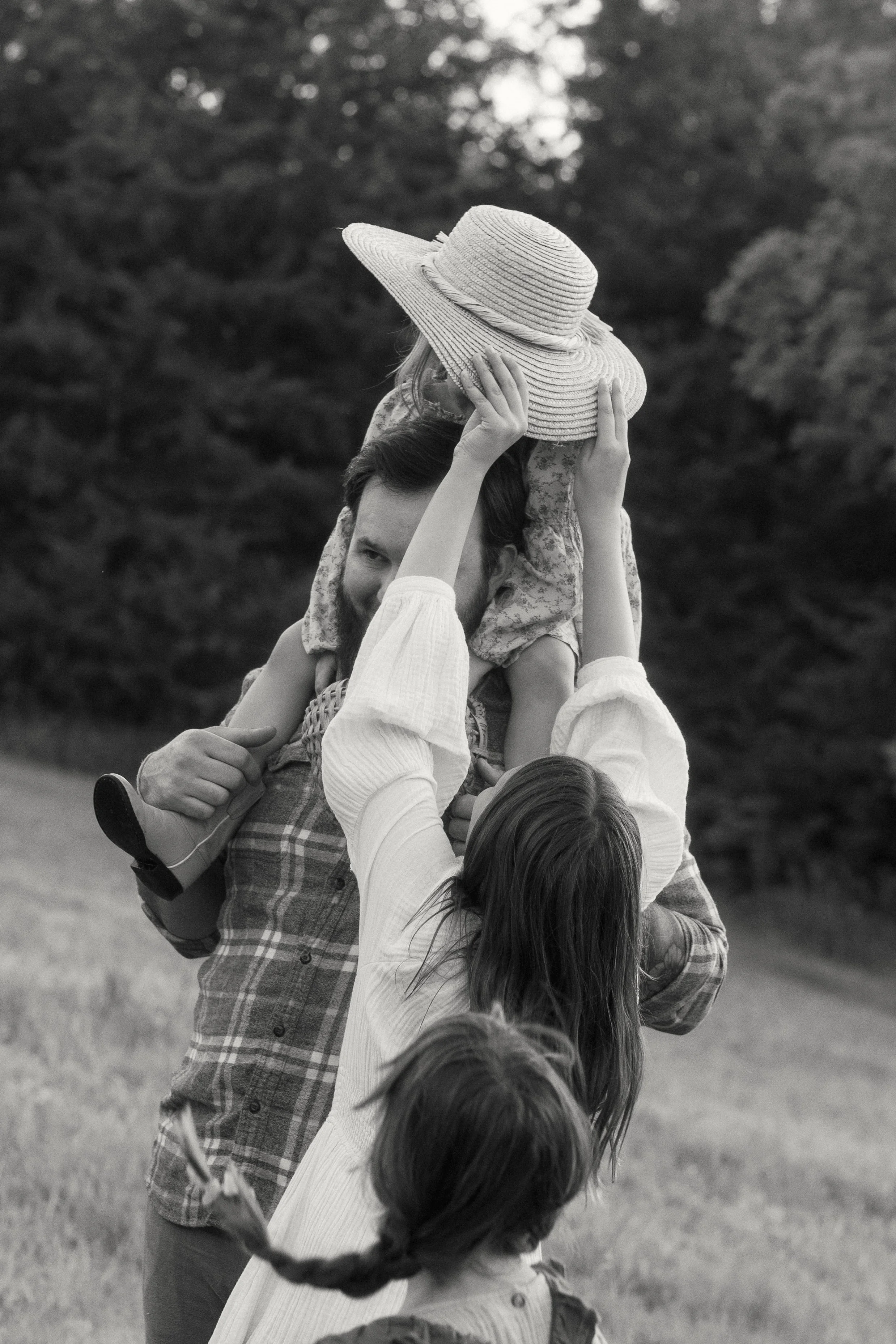 A family of four outdoors, with a man carrying a young girl on his shoulders, holding her hat, and a woman reaching up to touch the girl, with another child in front, all against a background of trees.