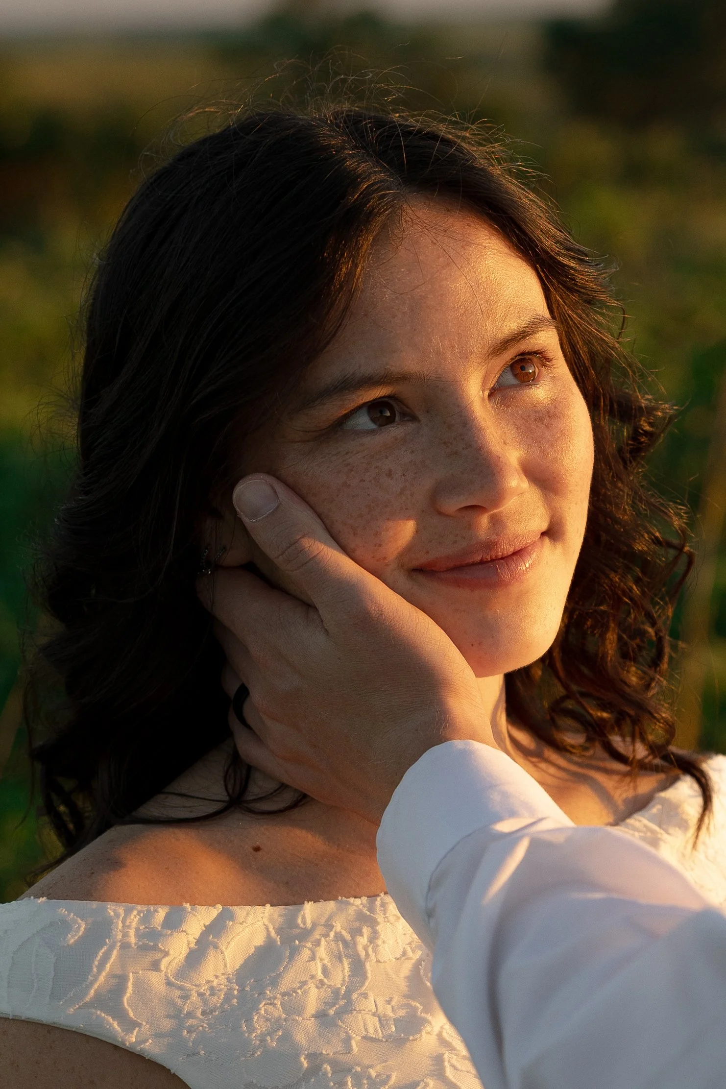 Close-up of a young woman with curly brown hair and brown eyes, resting her face on her hand, outdoors during golden hour, with blurred background and sunlight highlighting her features.