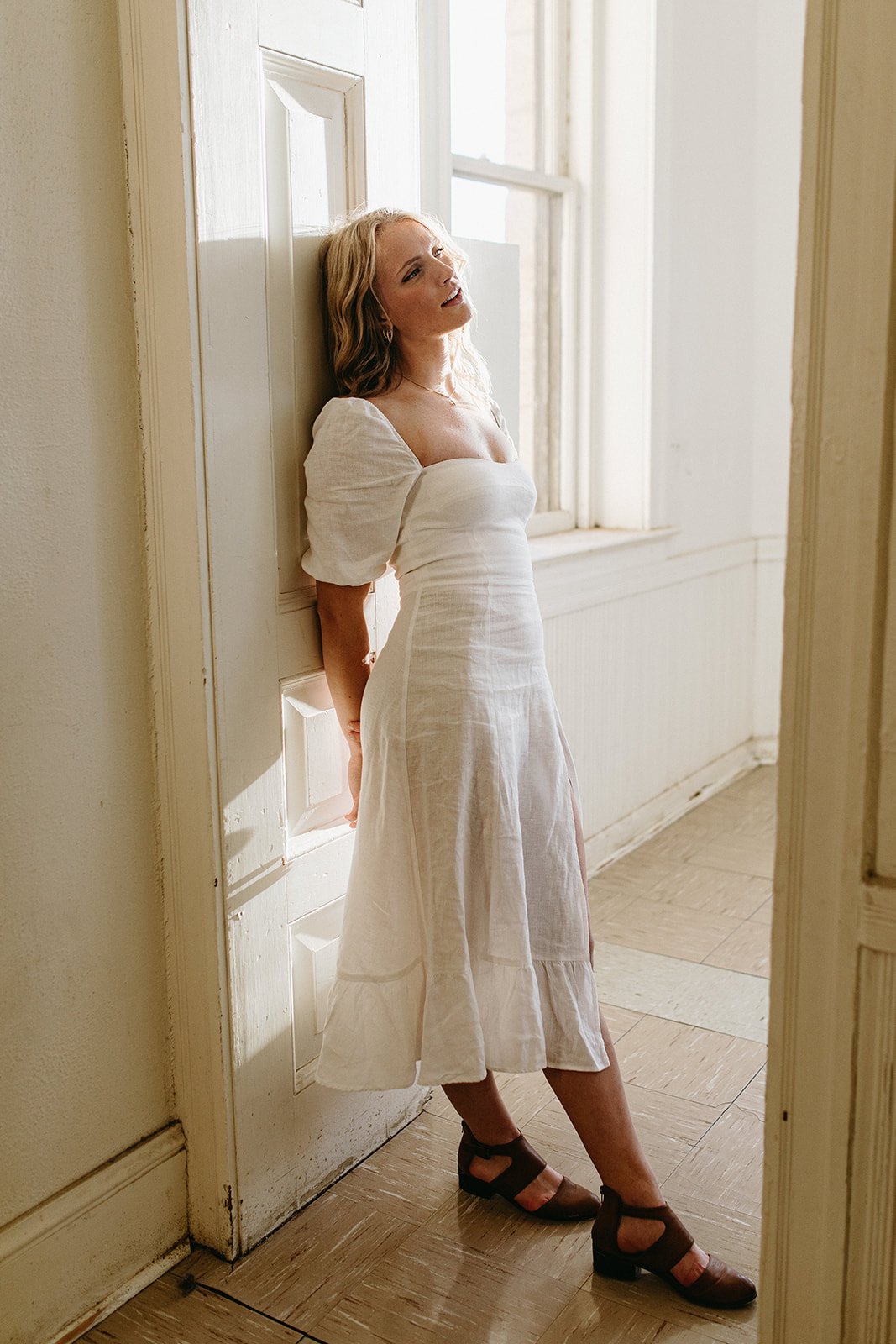 A woman in a white dress leaning against a doorframe near a window, with sunlight streaming in.
