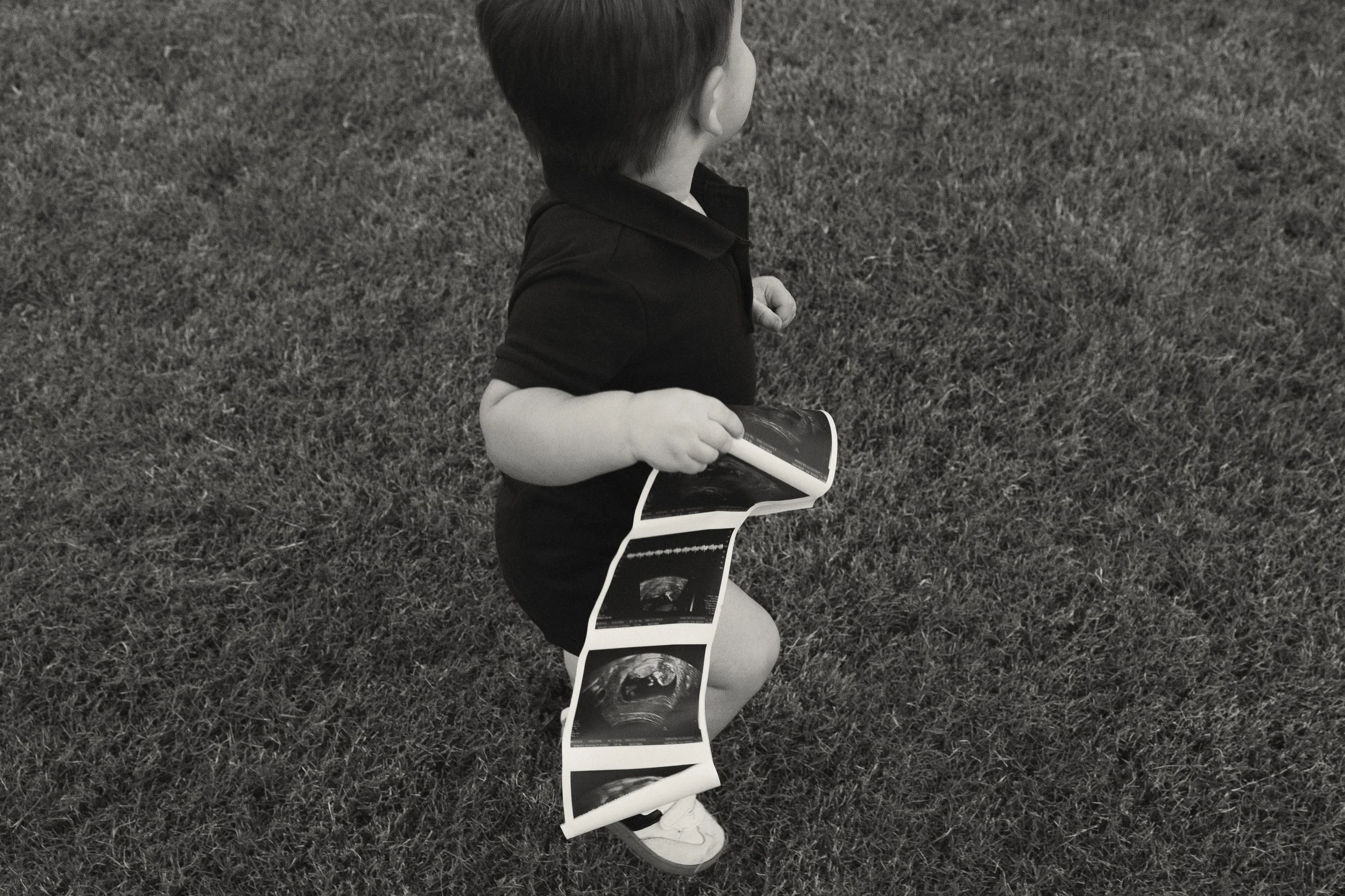 A young child kneels on grass, holding a strip of ultrasound photos.