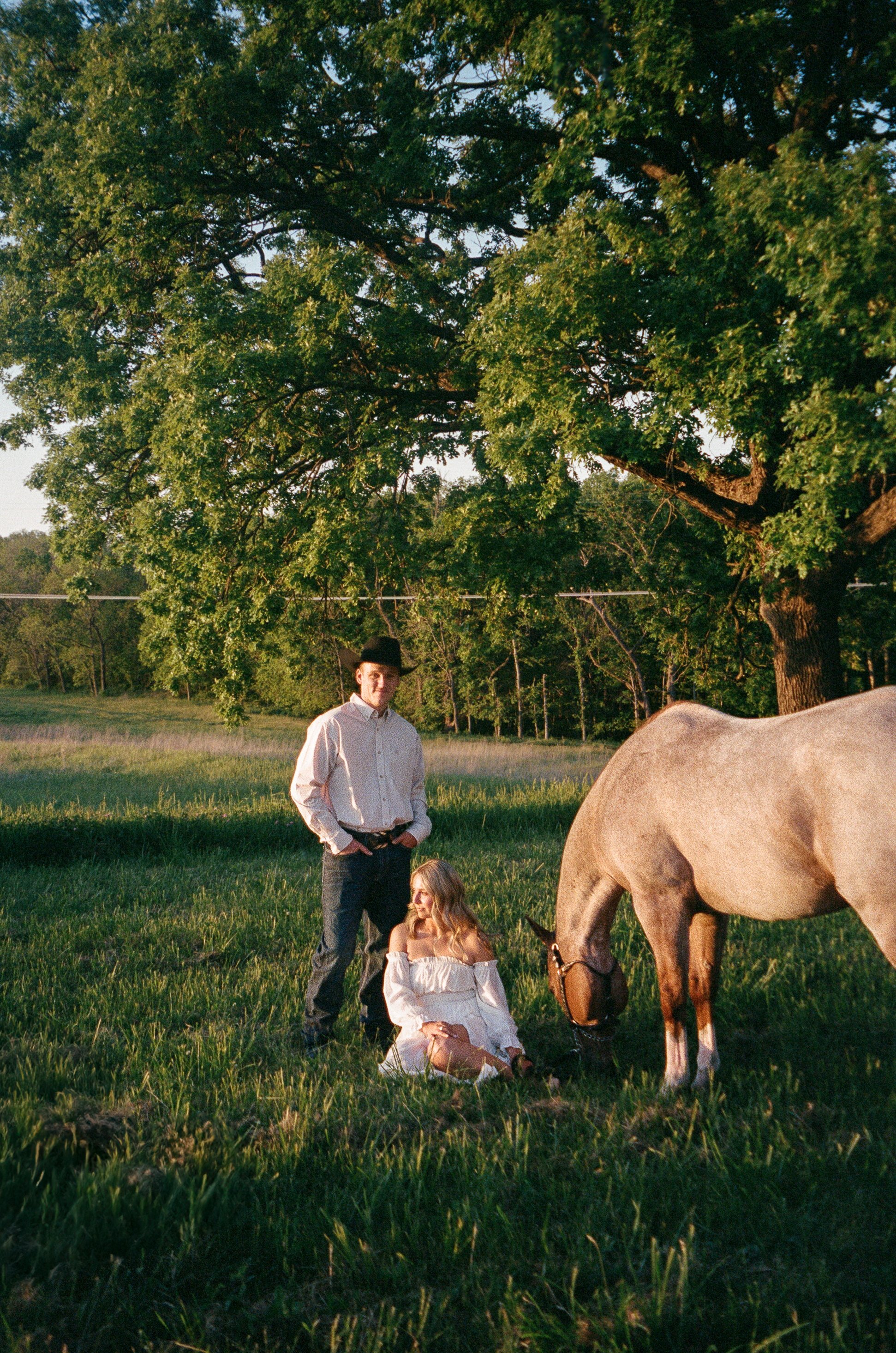 A young man in a white shirt and dark pants standing in a grassy field with a giant tree in the background. There is a woman sitting on the ground near a horse, which is grazing. The scene appears to be outdoors during sunset.