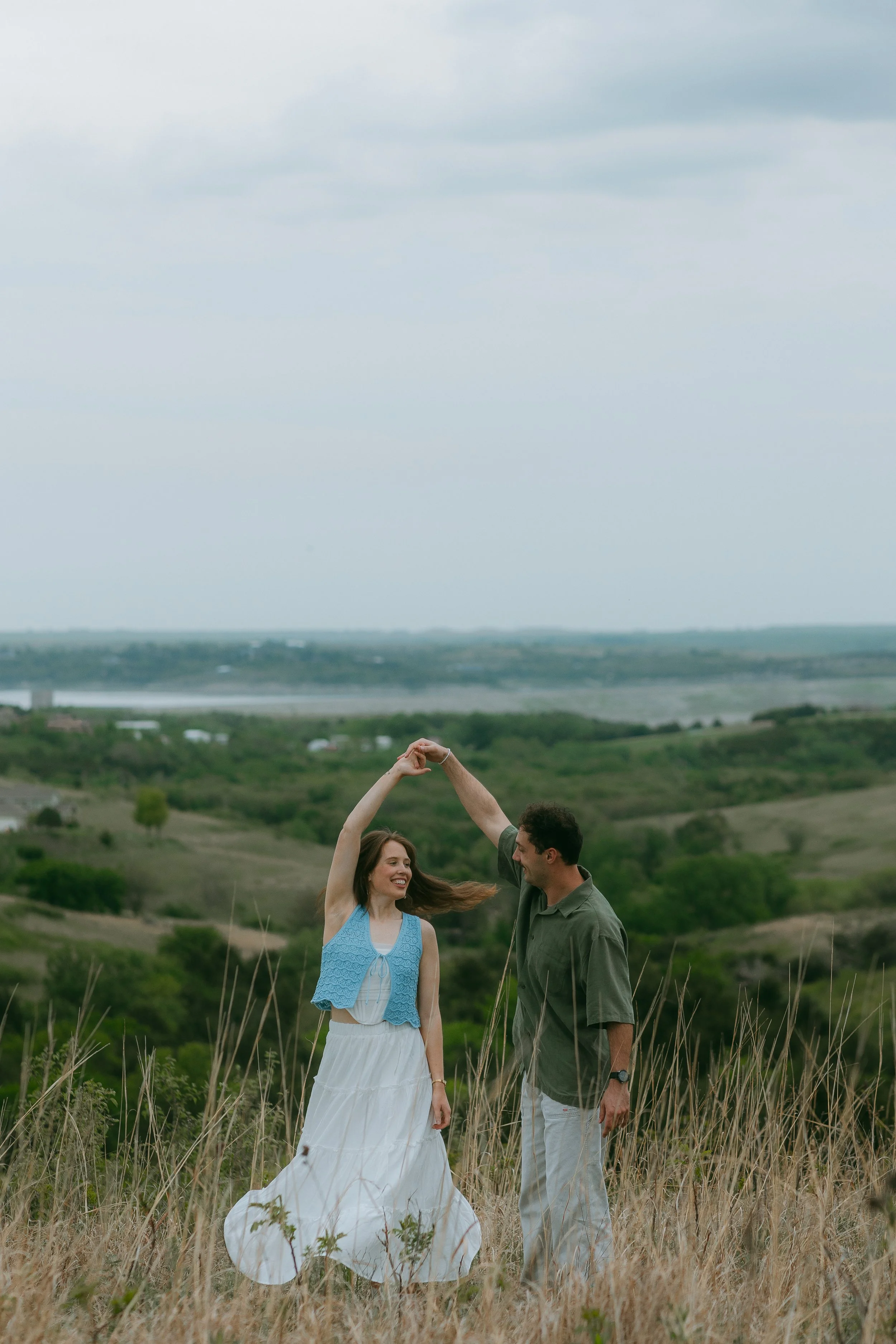 A couple dancing in a field with grass, with a scenic landscape of rolling hills and a body of water in the background on a cloudy day.