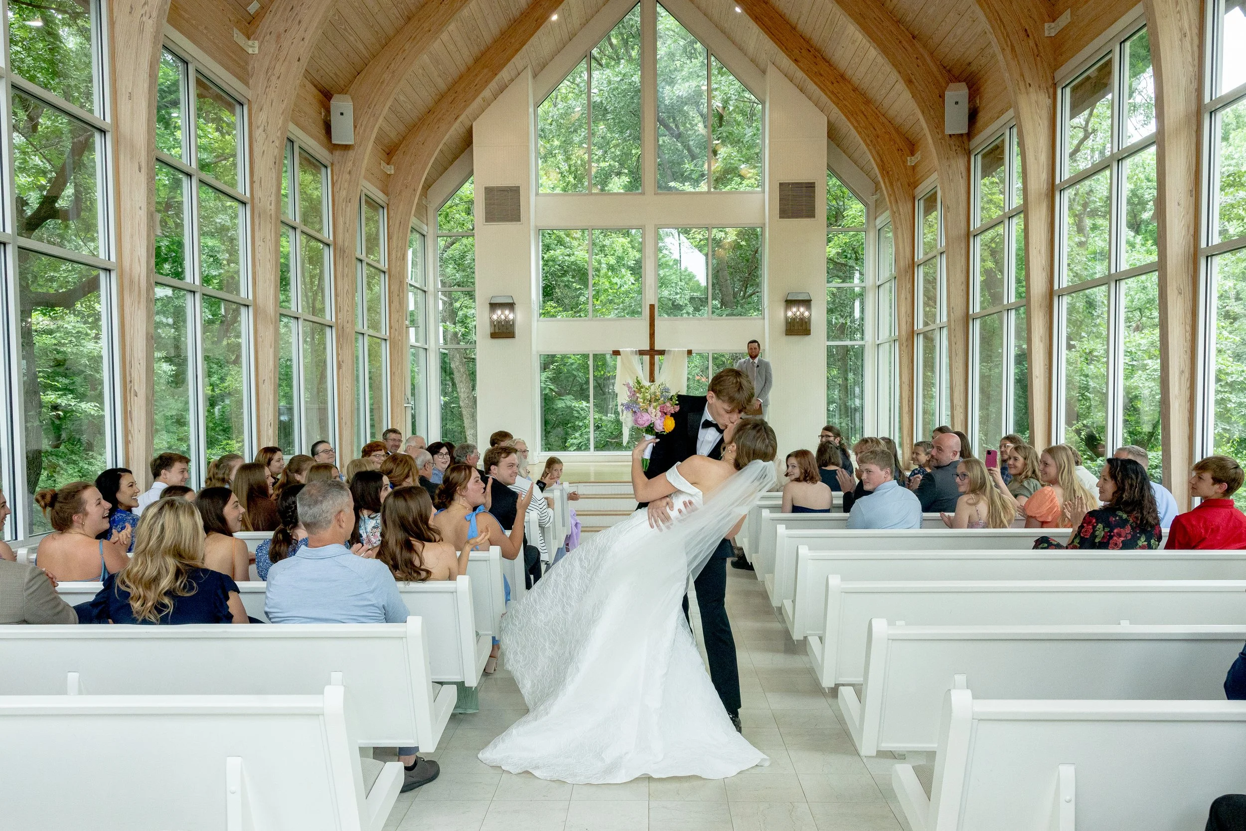 A wedding ceremony taking place inside a bright, modern church with large windows and wooden vaulted ceilings. The bride and groom are kissing at the altar, surrounded by seated guests who are smiling and taking photos.