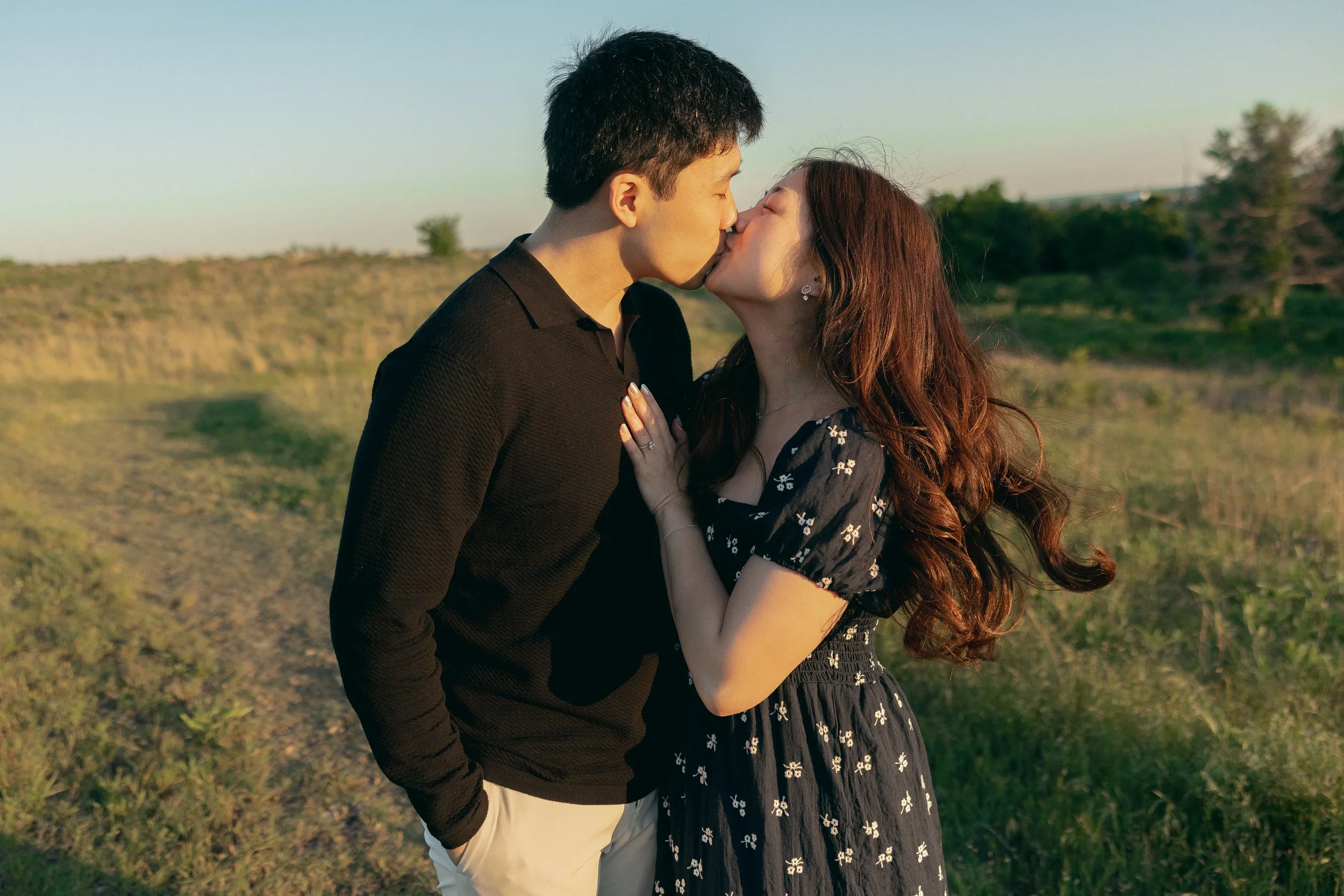 A couple kissing outdoors during sunset, standing in a field with trees in the background.