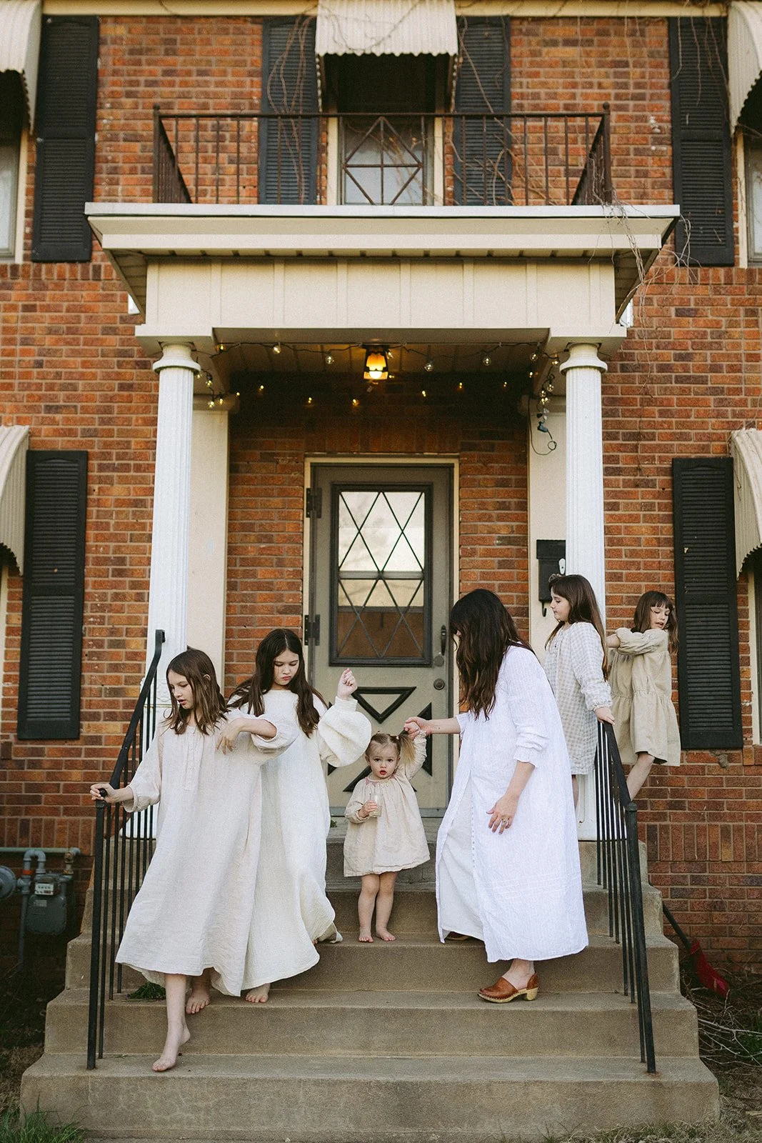 Six women and girls with light skin on a brick front porch, some touching or holding hands, others standing or sitting, dressed in neutral-colored dresses, with a house entrance in the background.