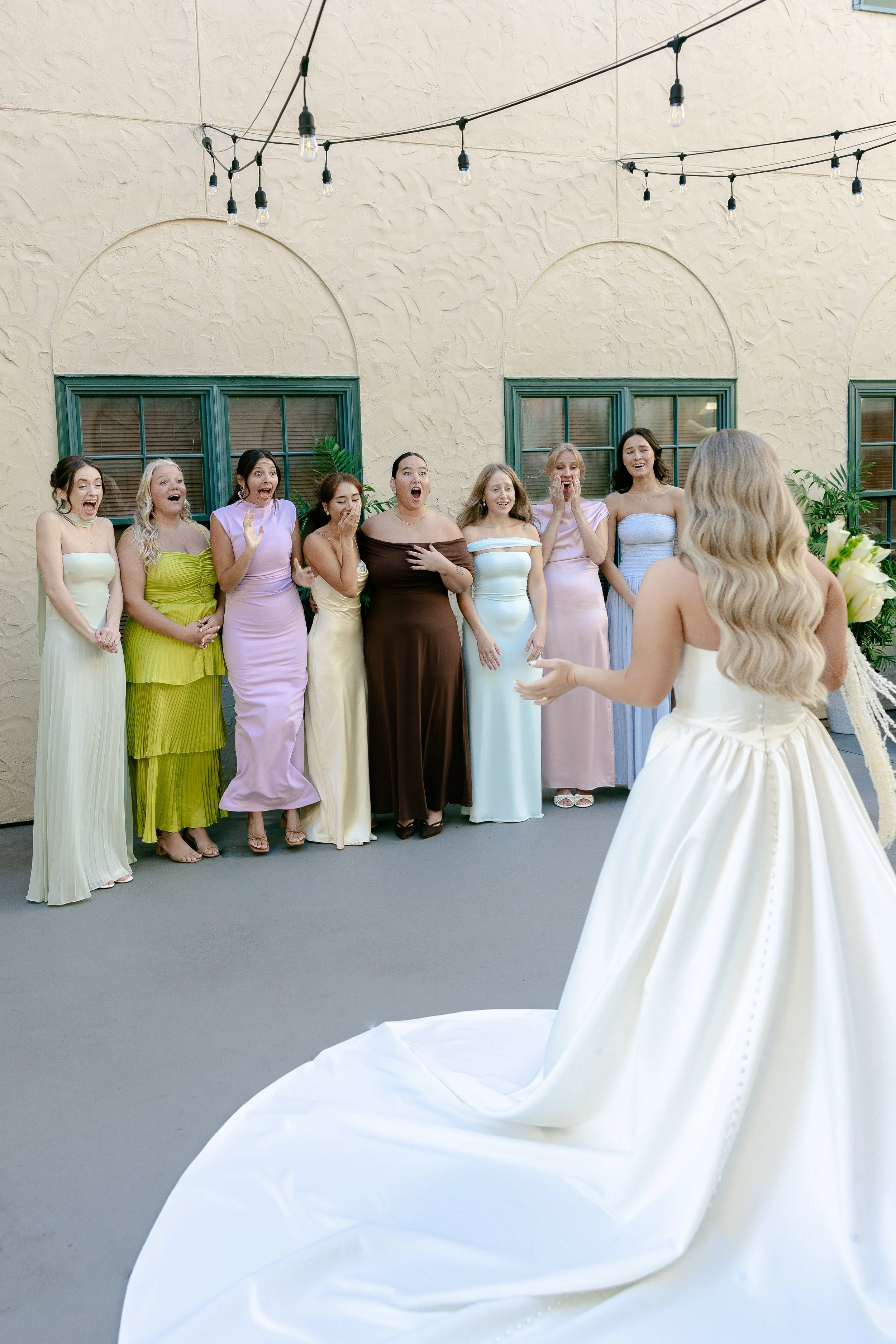 A bride in a white wedding dress facing a group of women, who are reacting with surprise and joy.