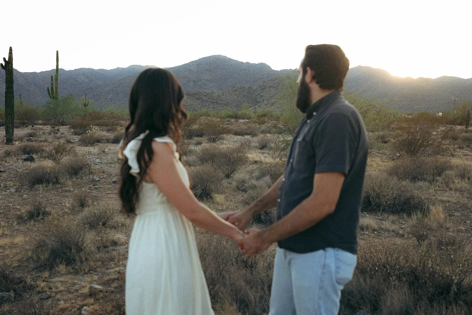 A couple holding hands and gazing at each other in a desert landscape with mountains and cacti at sunset.