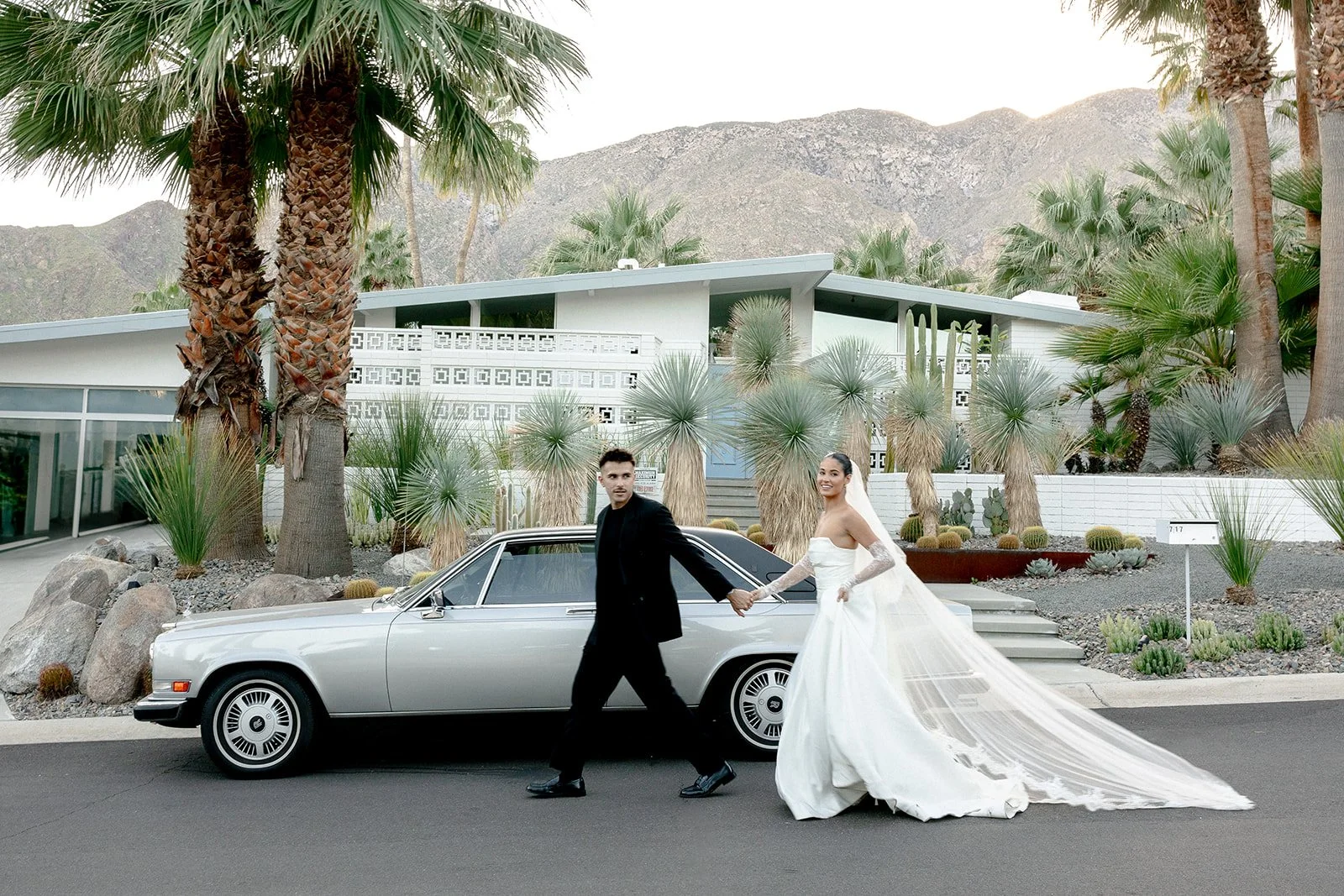 A bride and groom holding hands and walking in front of a vintage car on a residential street in front of a modern house with desert landscaping, palm trees, and mountains in the background.