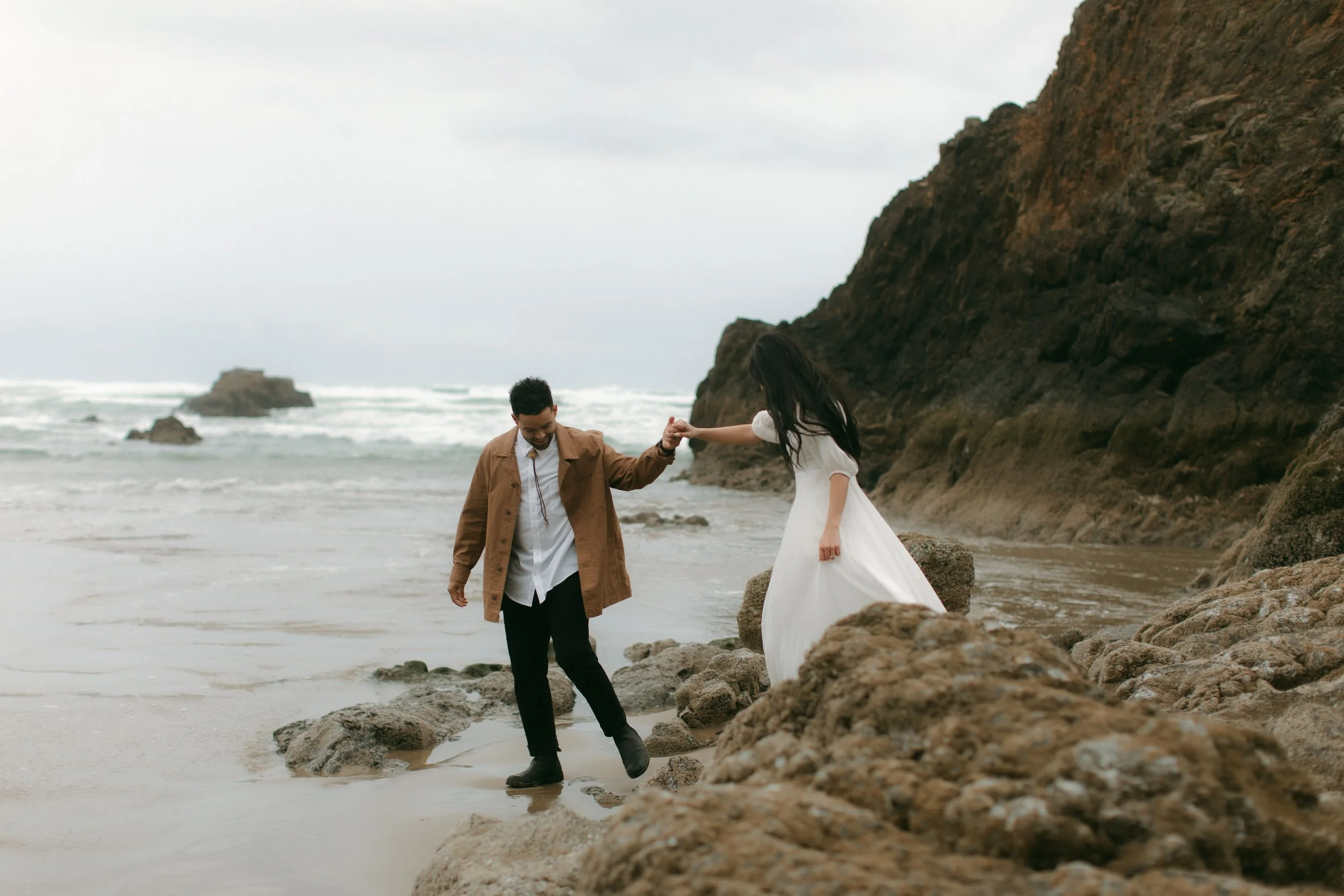 A couple walking on a rocky beach, with the man helping the woman, who is wearing a white dress, navigate over the rocks near the water.