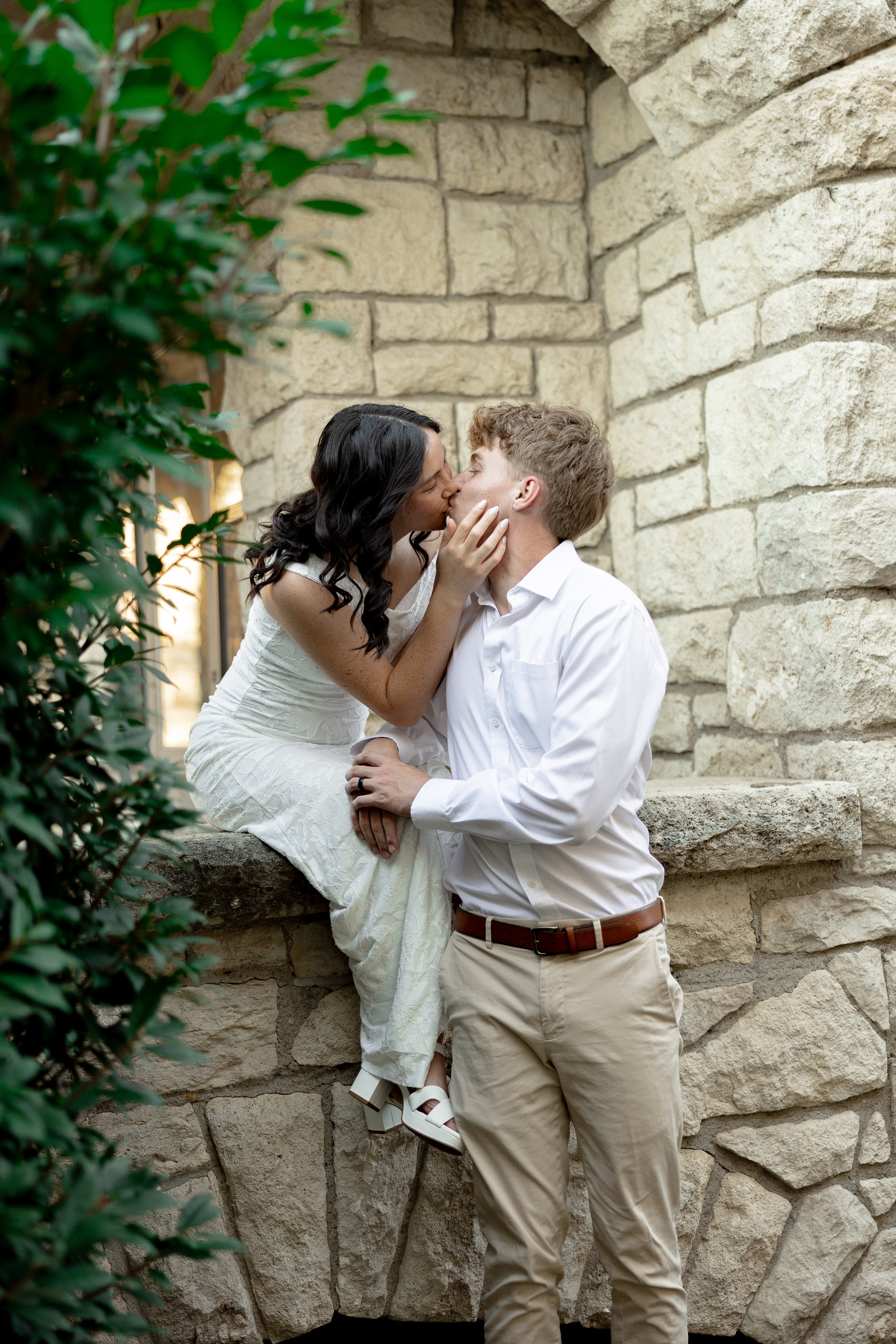 A couple kissing outdoors, the woman sitting on a stone ledge and the man standing in front of her, with a stone wall background and green foliage on the left side.